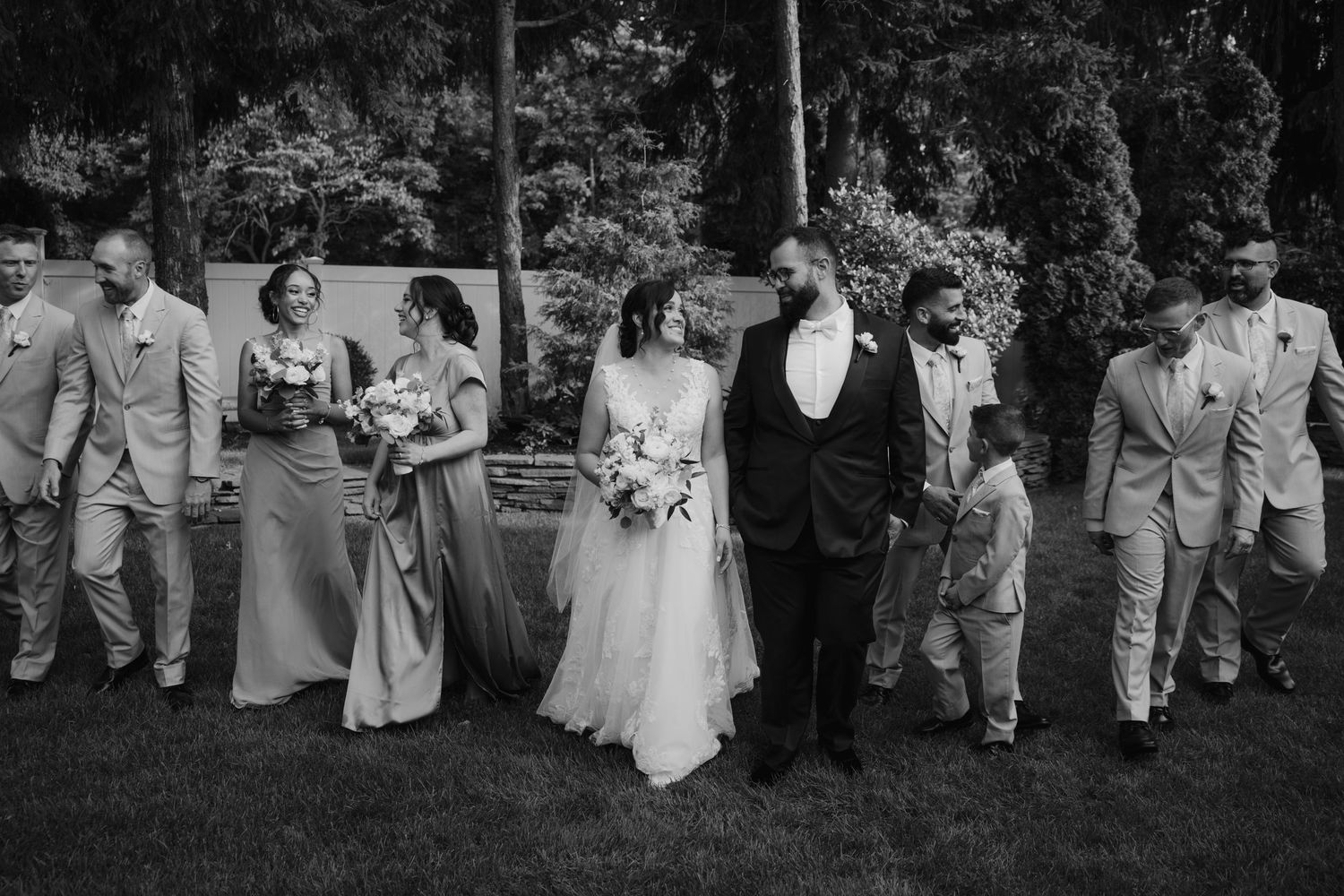 Black and white wedding party photo with bride, groom, bridesmaids, and groomsmen walking outdoors.