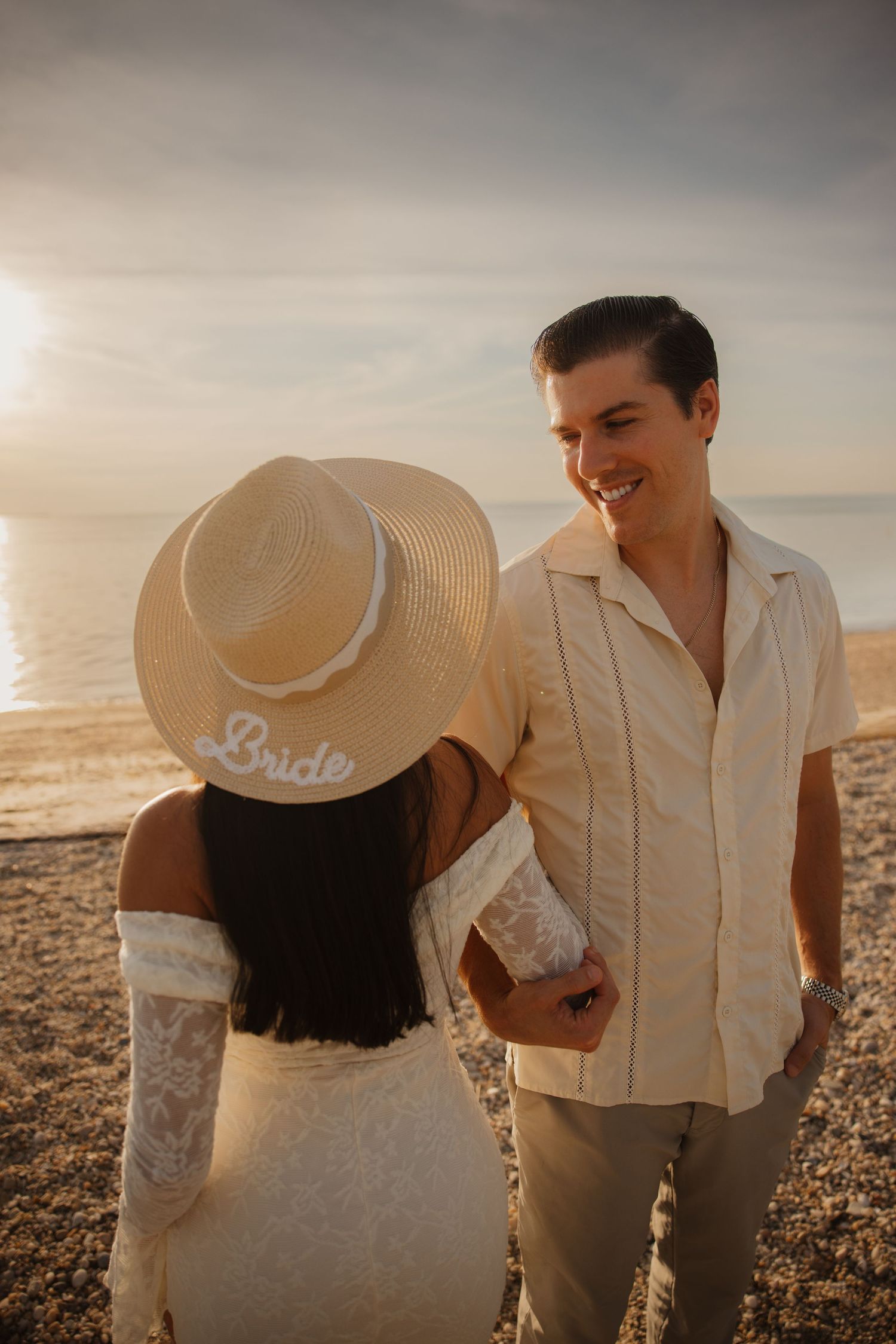Couple holding hands on a beach at sunset, bride wearing a straw hat and lace dress, groom smiling in cream shirt.