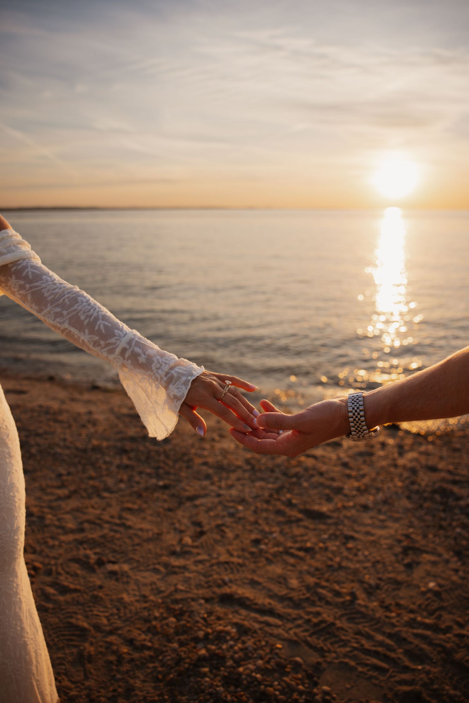 Two people holding hands on a sandy beach at sunset, one wearing a white lace dress and silver bracelet.