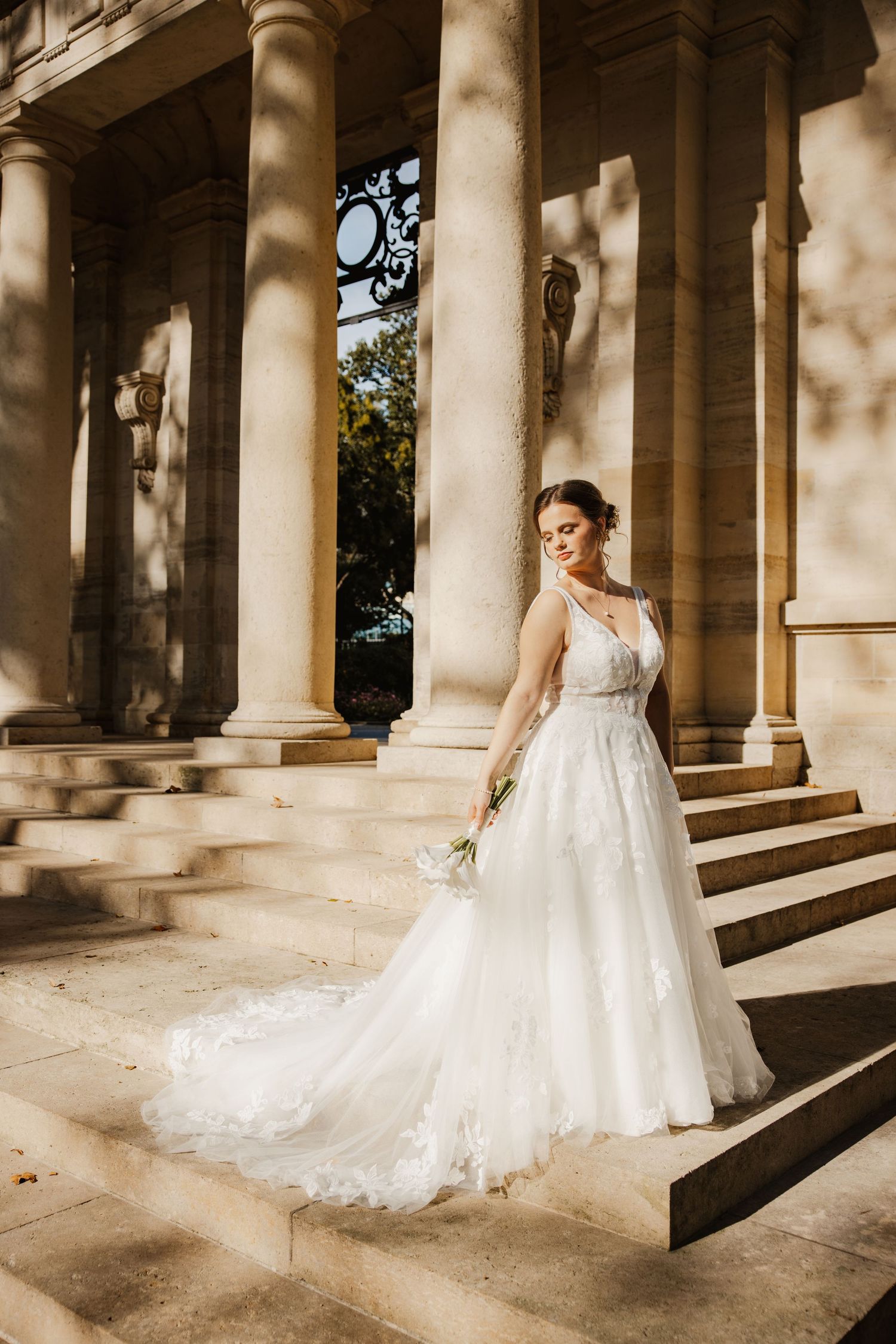 Bride in white lace gown with train poses on stone steps between elegant columns holding a bouquet.