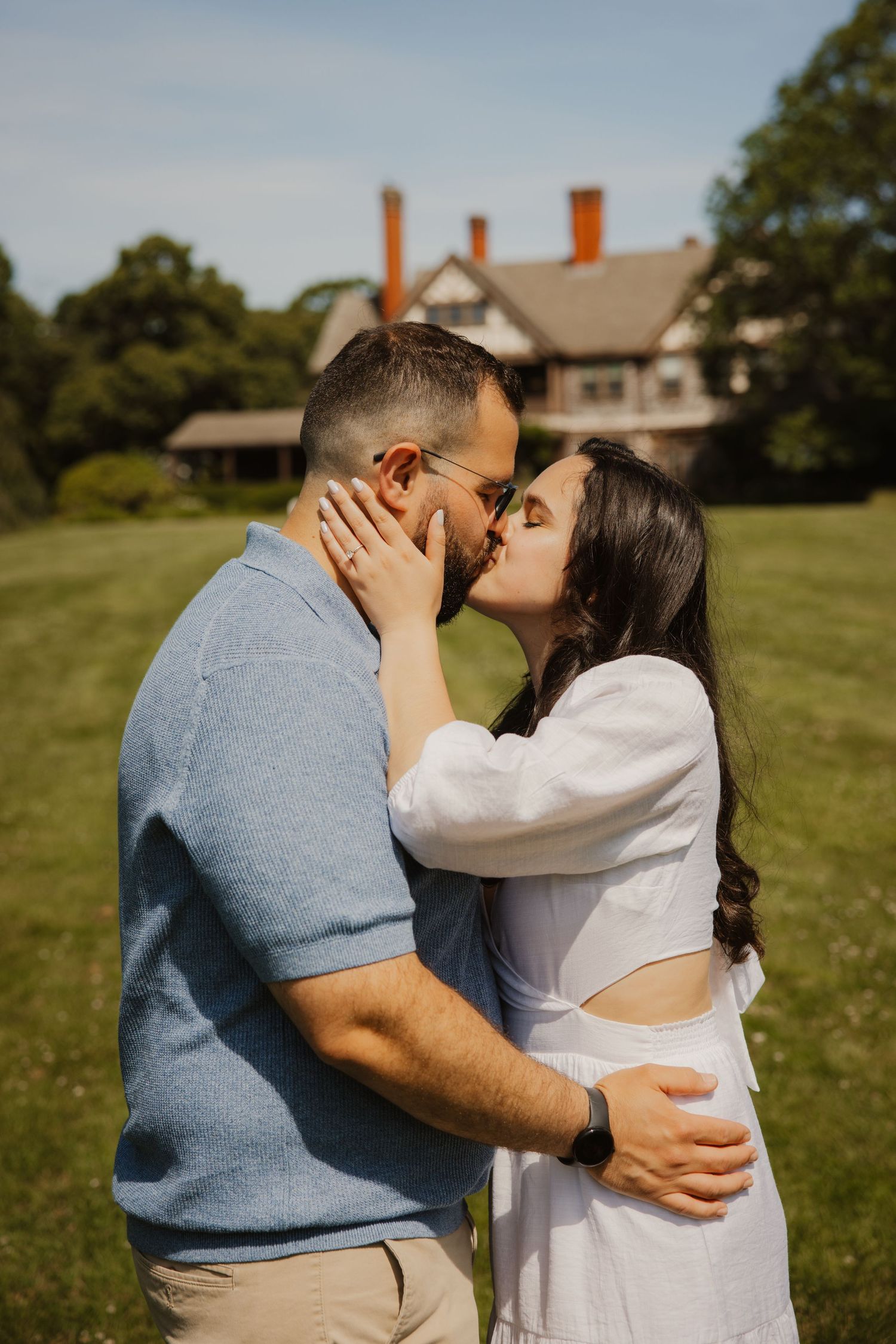 A couple shares a romantic kiss on a green lawn with a charming brick estate visible behind them.
