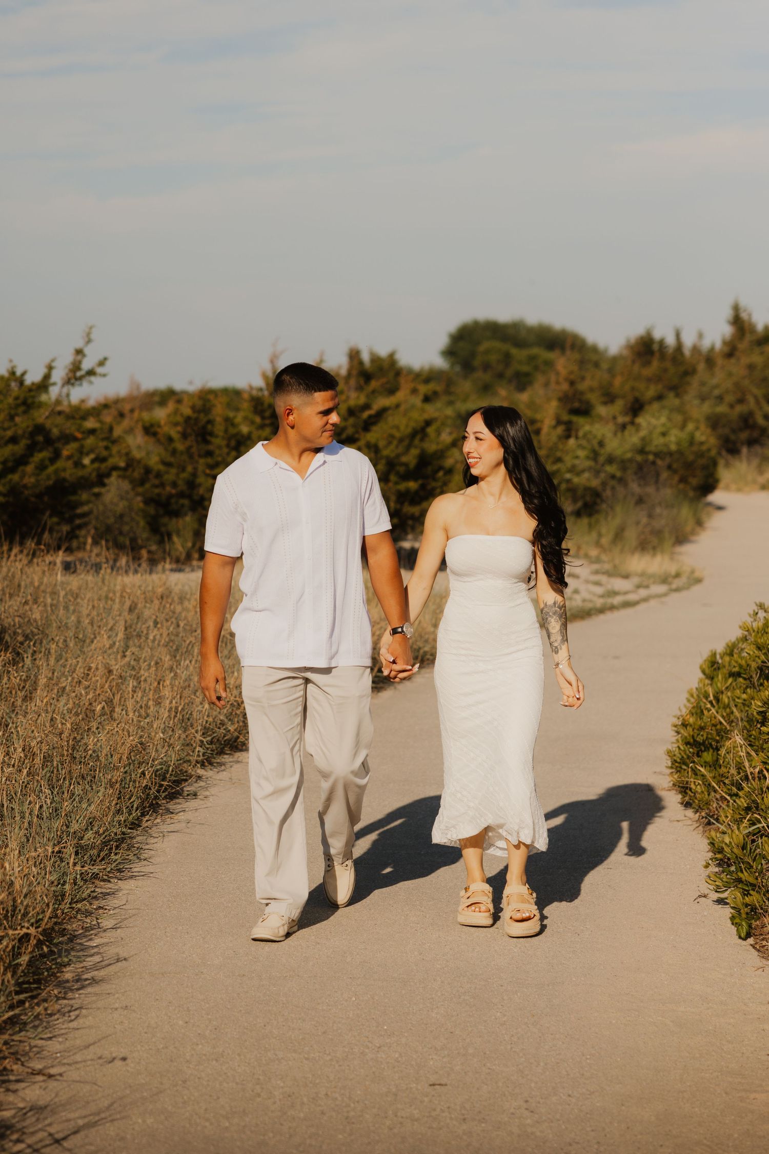 Happy couple holding hands and walking along sunny sandy path surrounded by coastal shrubs.