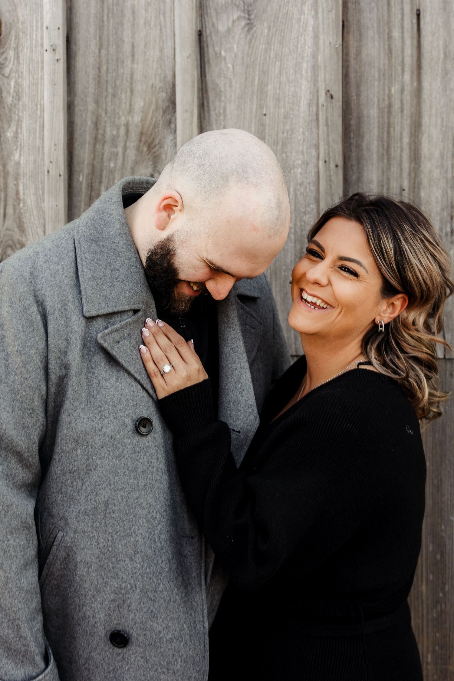 Laughing woman in black shows engagement ring while leaning against bald bearded man in gray coat.