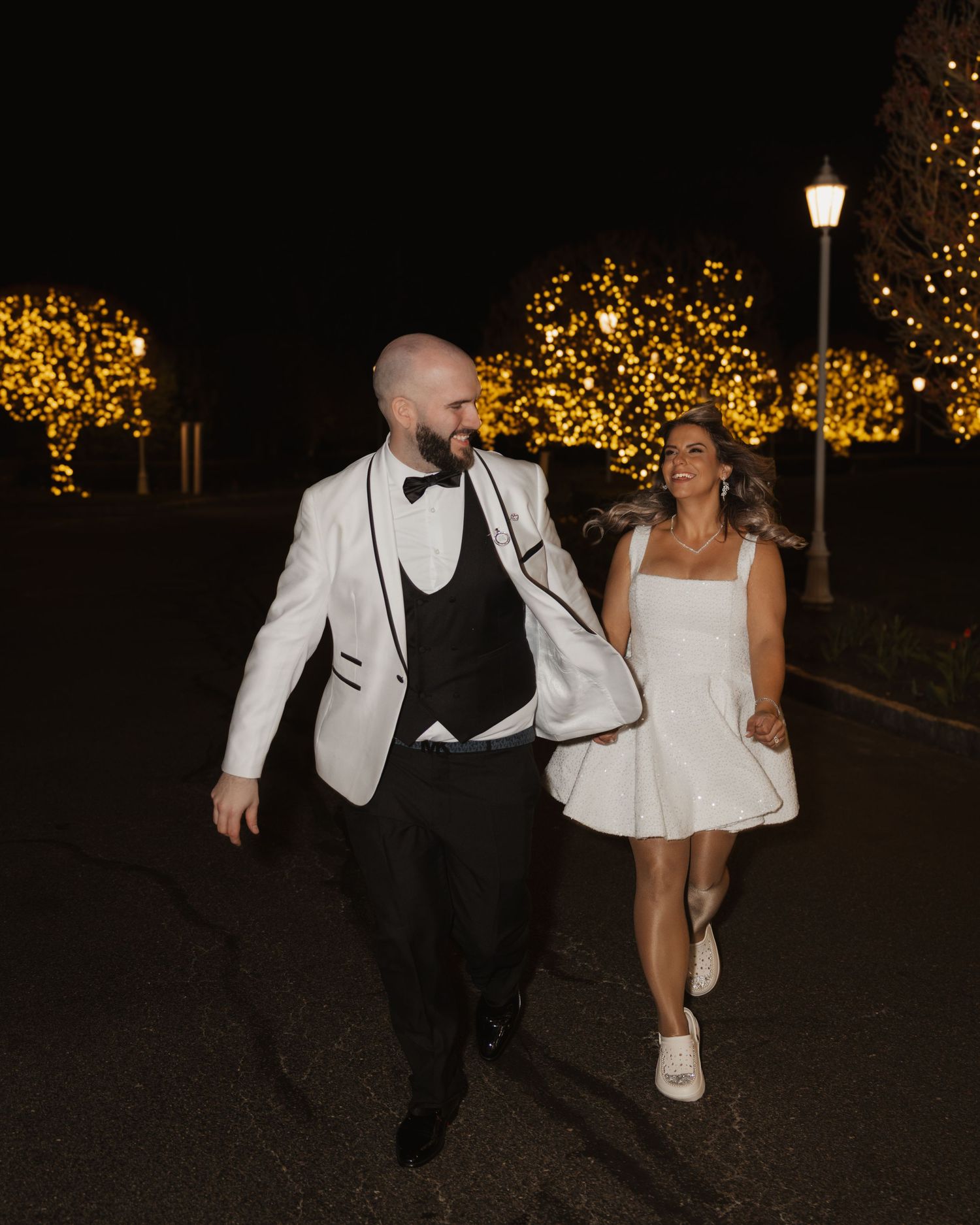Couple in wedding attire walking hand in hand at night amid golden lit trees.
