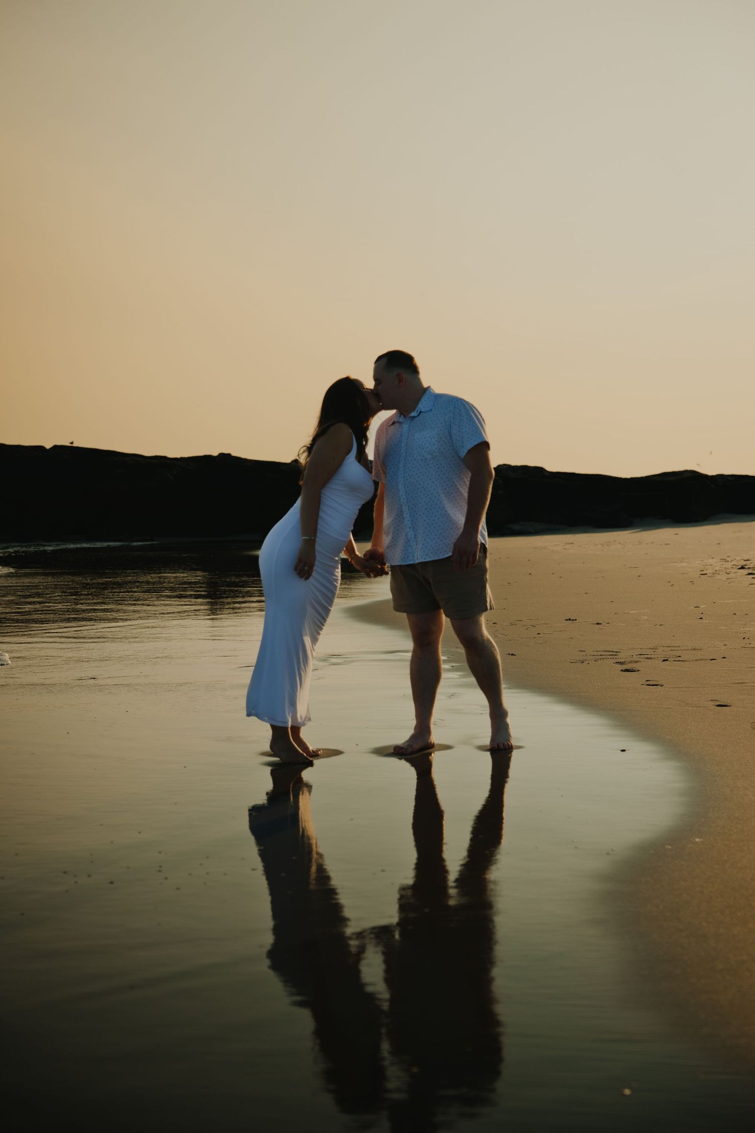 Silhouette of couple kissing on wet sand beach at sunset with reflections in water.