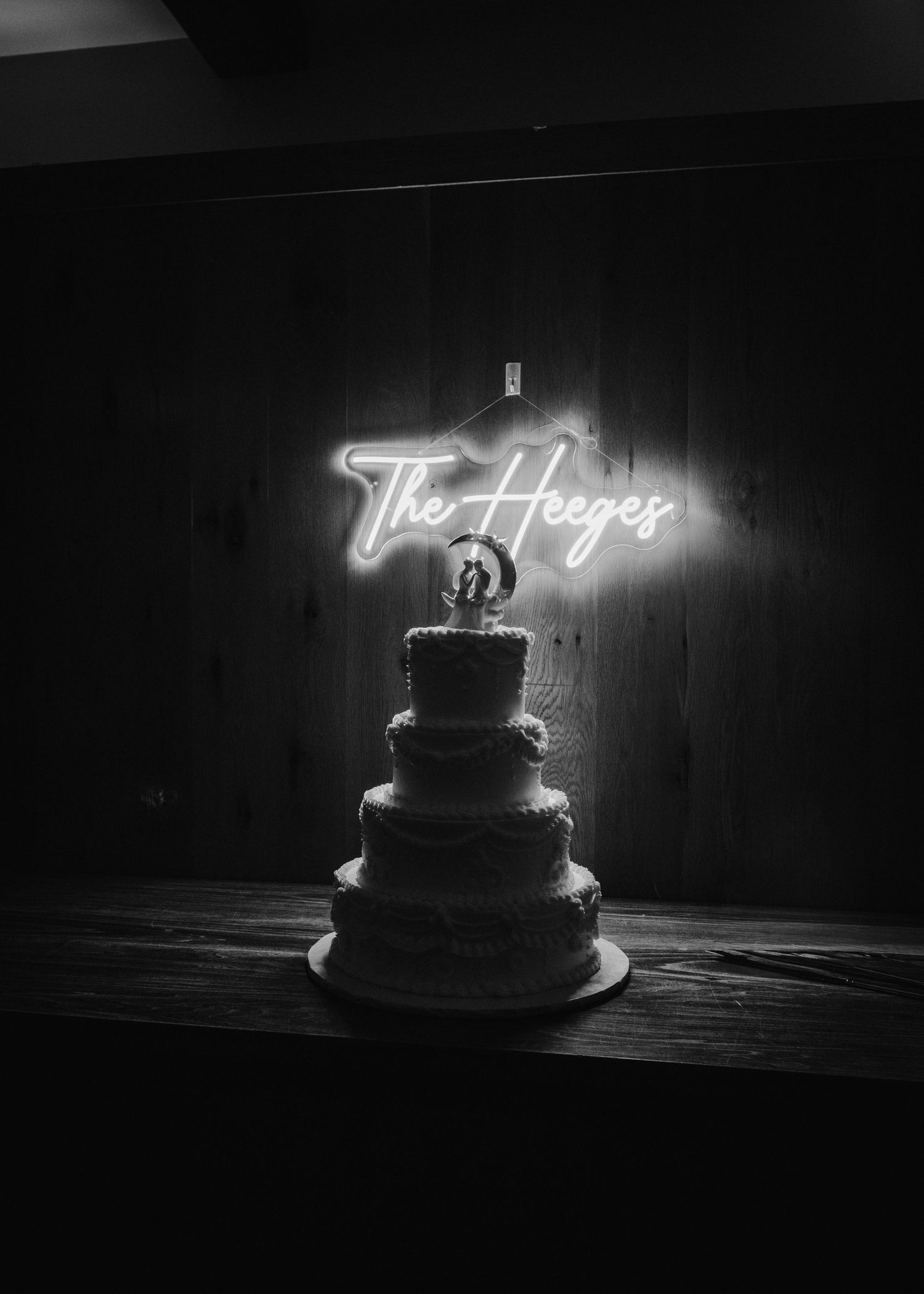 Four-tier wedding cake with topper glows beneath a neon sign reading The Heeges in dark room.