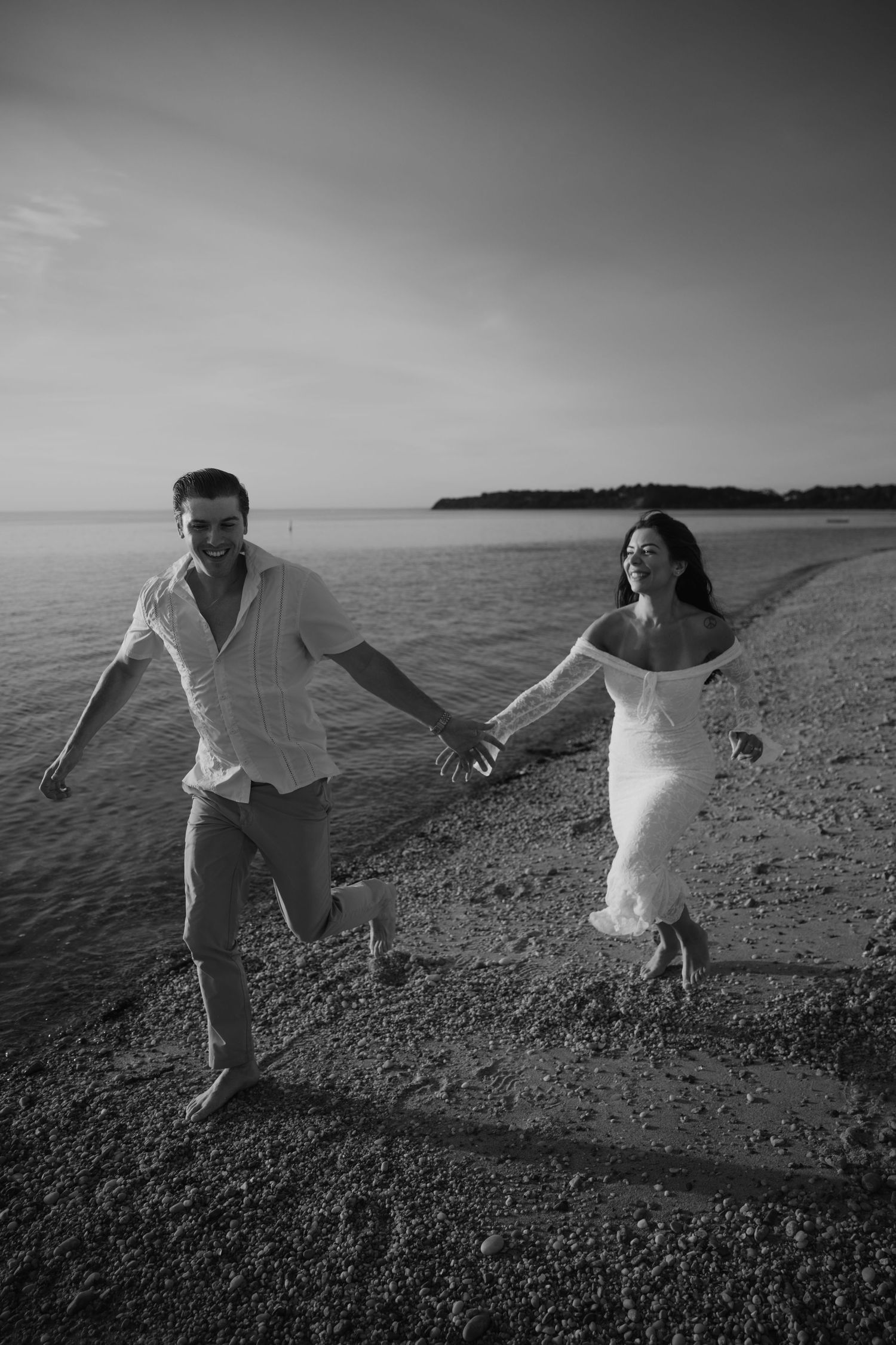 Black and white photo of a couple holding hands and running joyfully along a sandy beach at sunset.