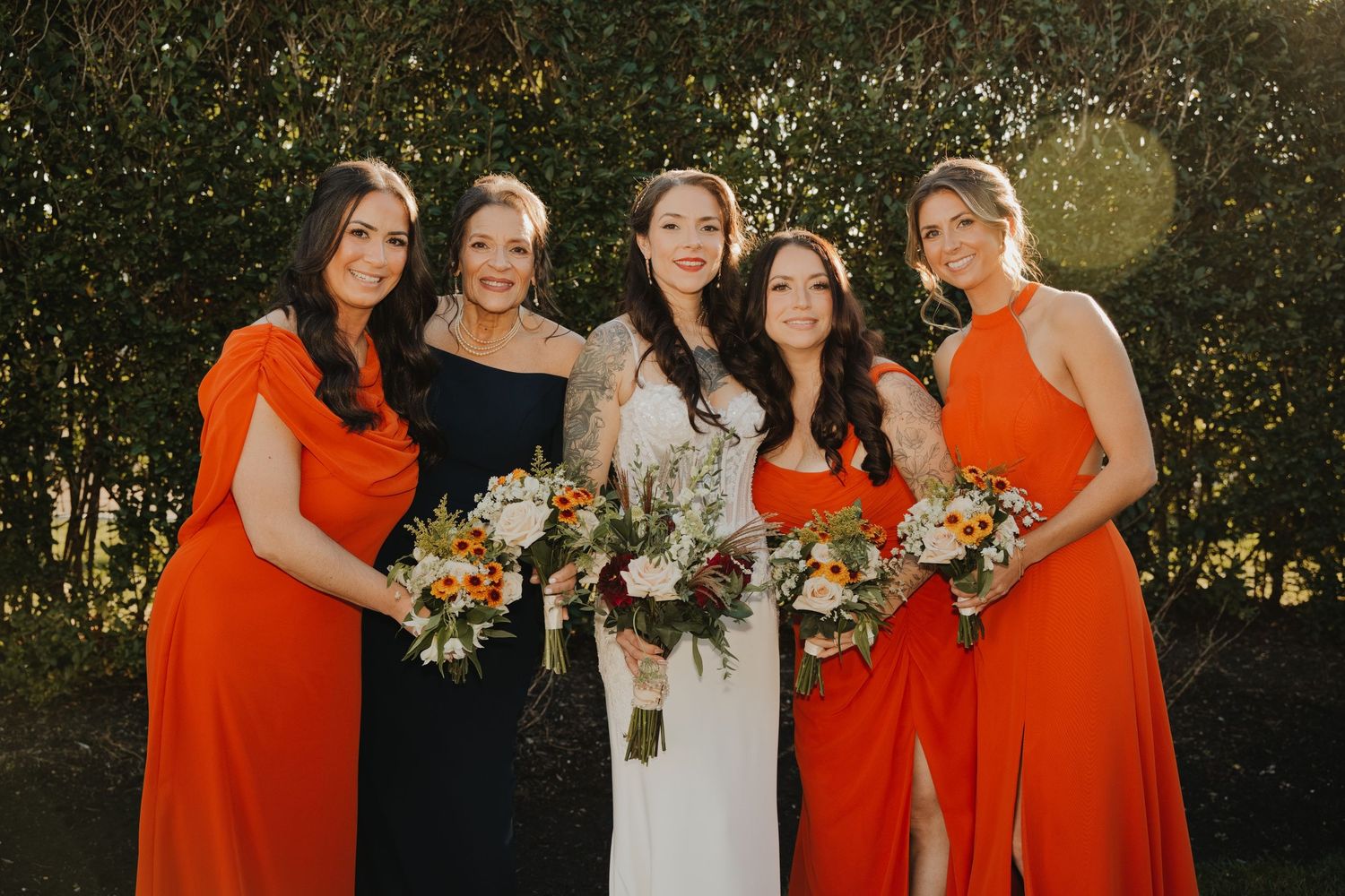 Bride in white gown poses with four bridesmaids in orange and black dresses, each holding a colorful floral bouquet outdoors.