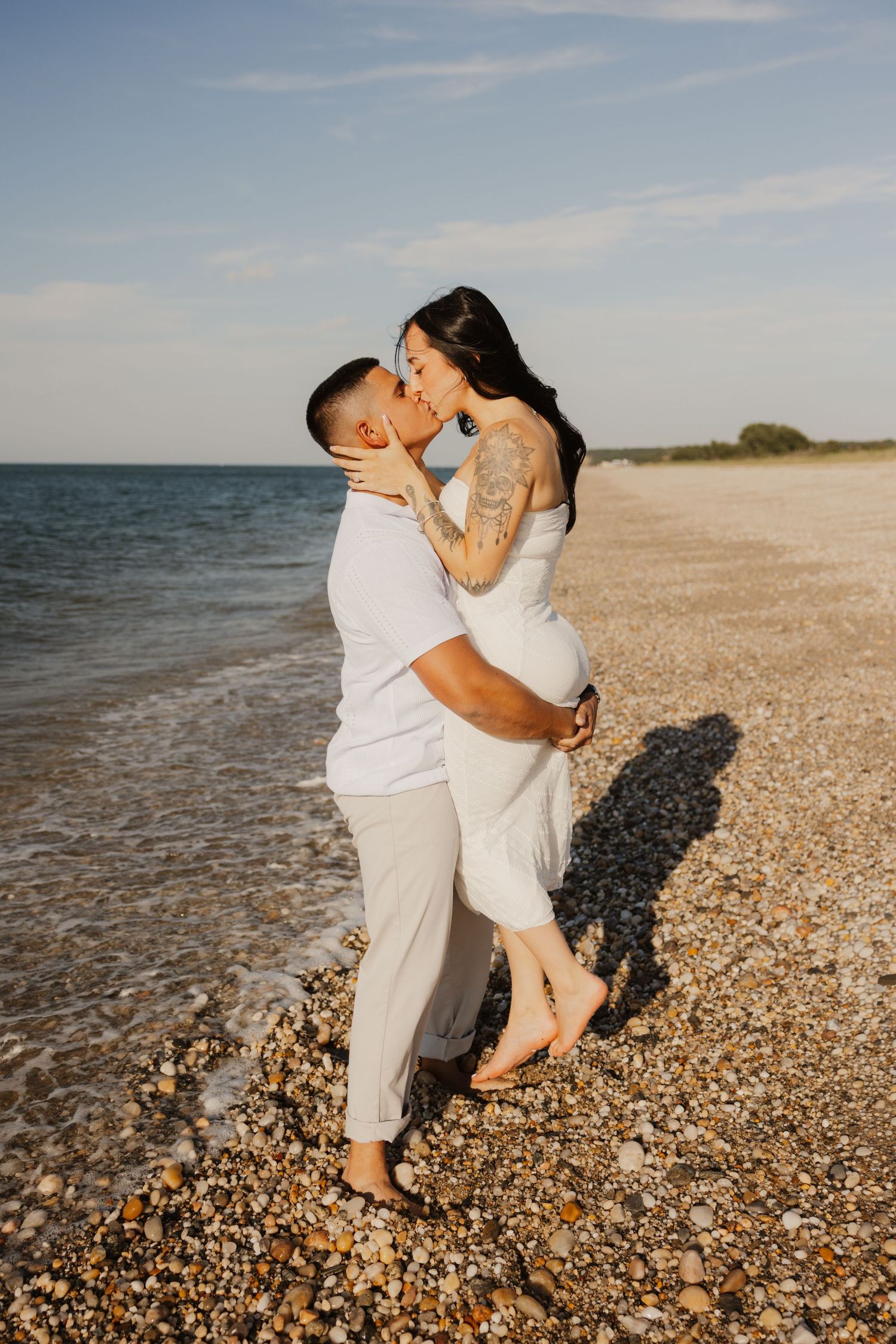 Couple kissing on rocky beach shoreline at golden hour with calm ocean in background.