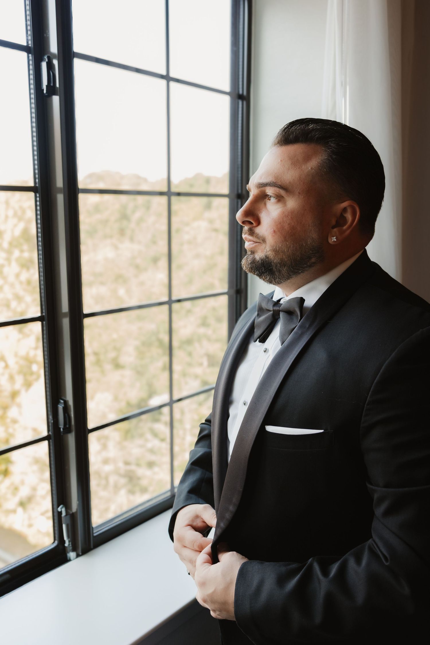 Dapper groom in black tuxedo with bow tie gazing thoughtfully out a window.