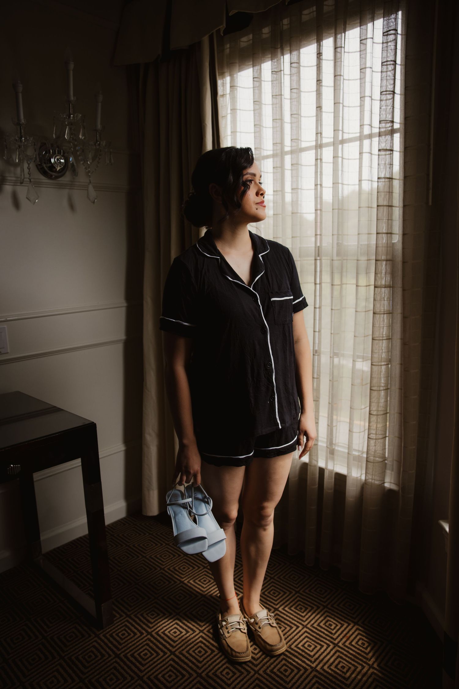 Bride in black pajamas stands by a sunlit window holding heels in a hotel room before wedding.