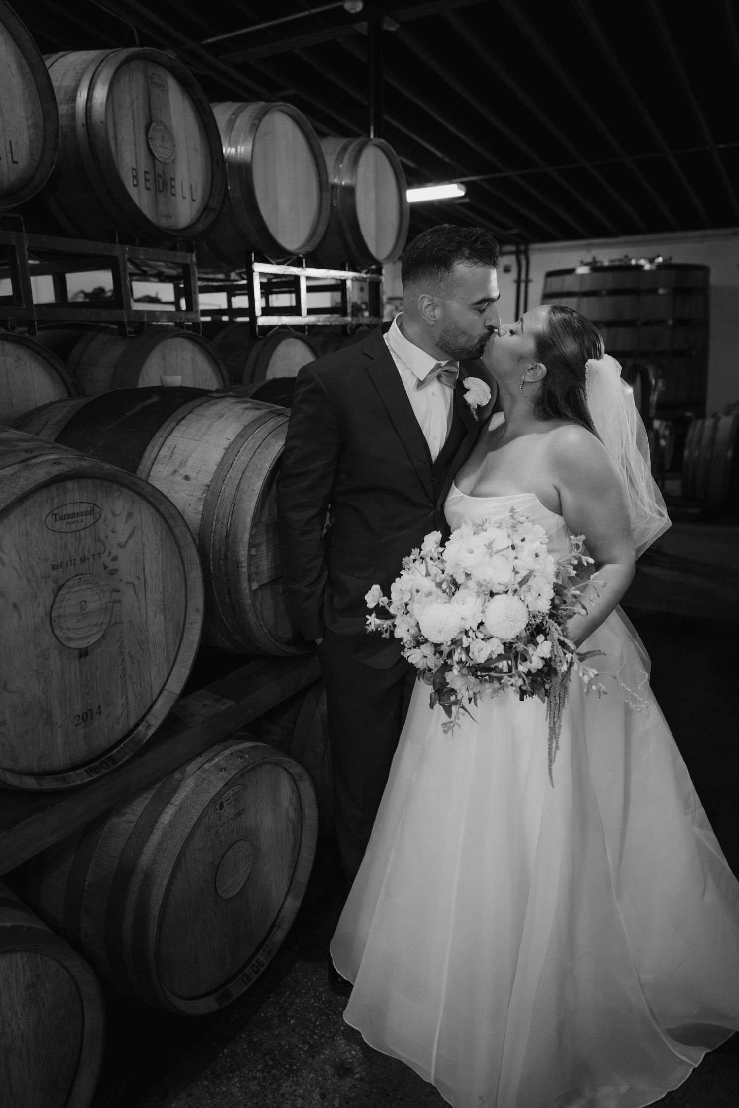 Black and white photo of a bride and groom kissing in a winery barrel room, bride holding a white floral bouquet.