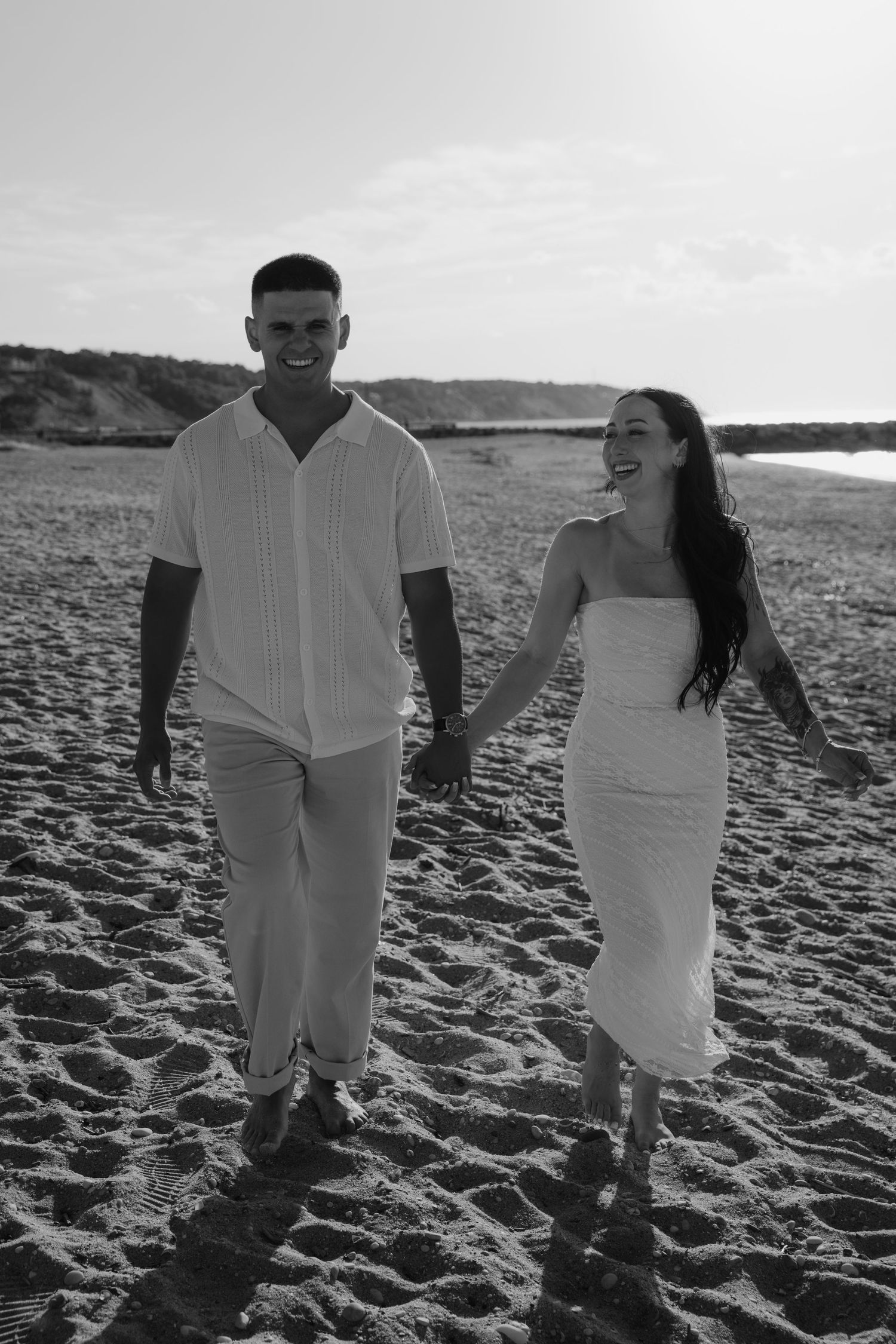 Black and white photo of a couple holding hands and walking barefoot on a sandy beach at sunset.