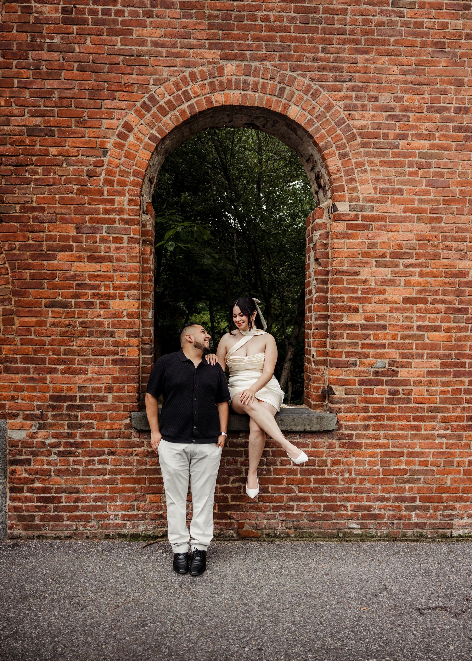 Couple posing at a brick archway, woman seated on ledge smiling at her standing partner.
