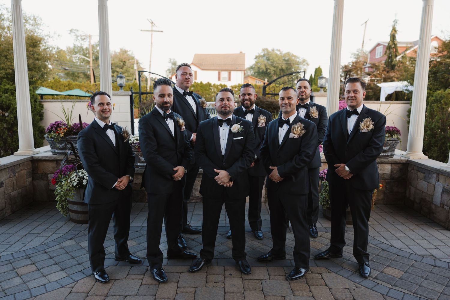 Groom and groomsmen posing together in matching black tuxedos at outdoor wedding venue.
