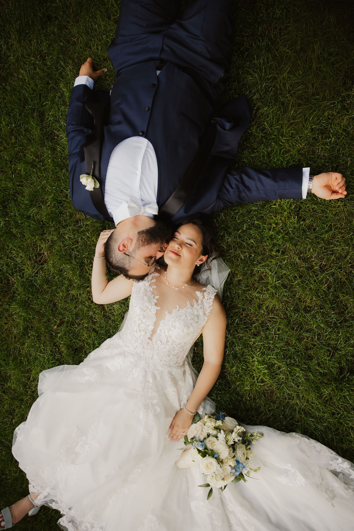 Bride and groom lie on lush green grass laughing together in a joyful wedding portrait.