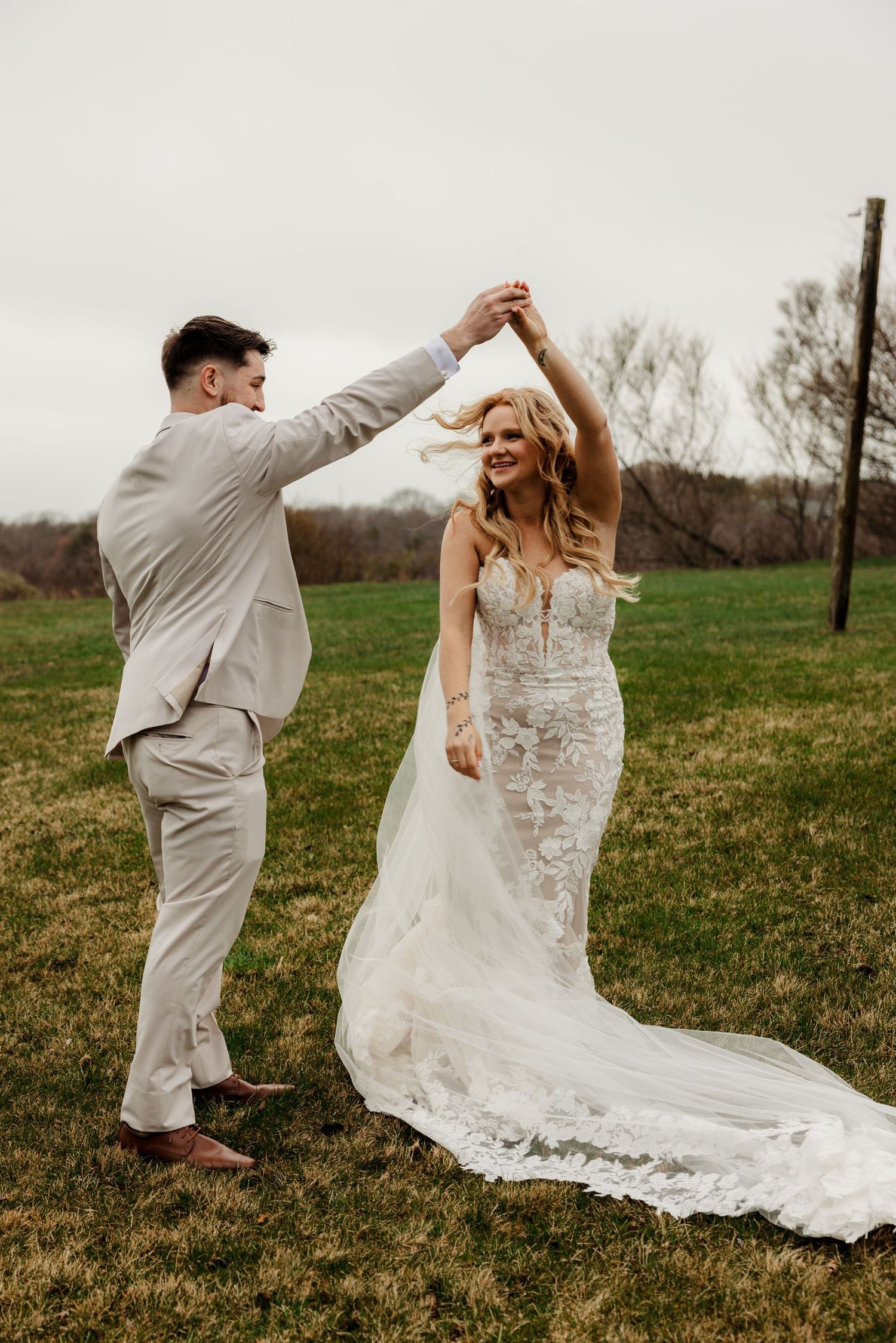 Groom twirling bride in floral wedding gown on a green outdoor field on cloudy day.
