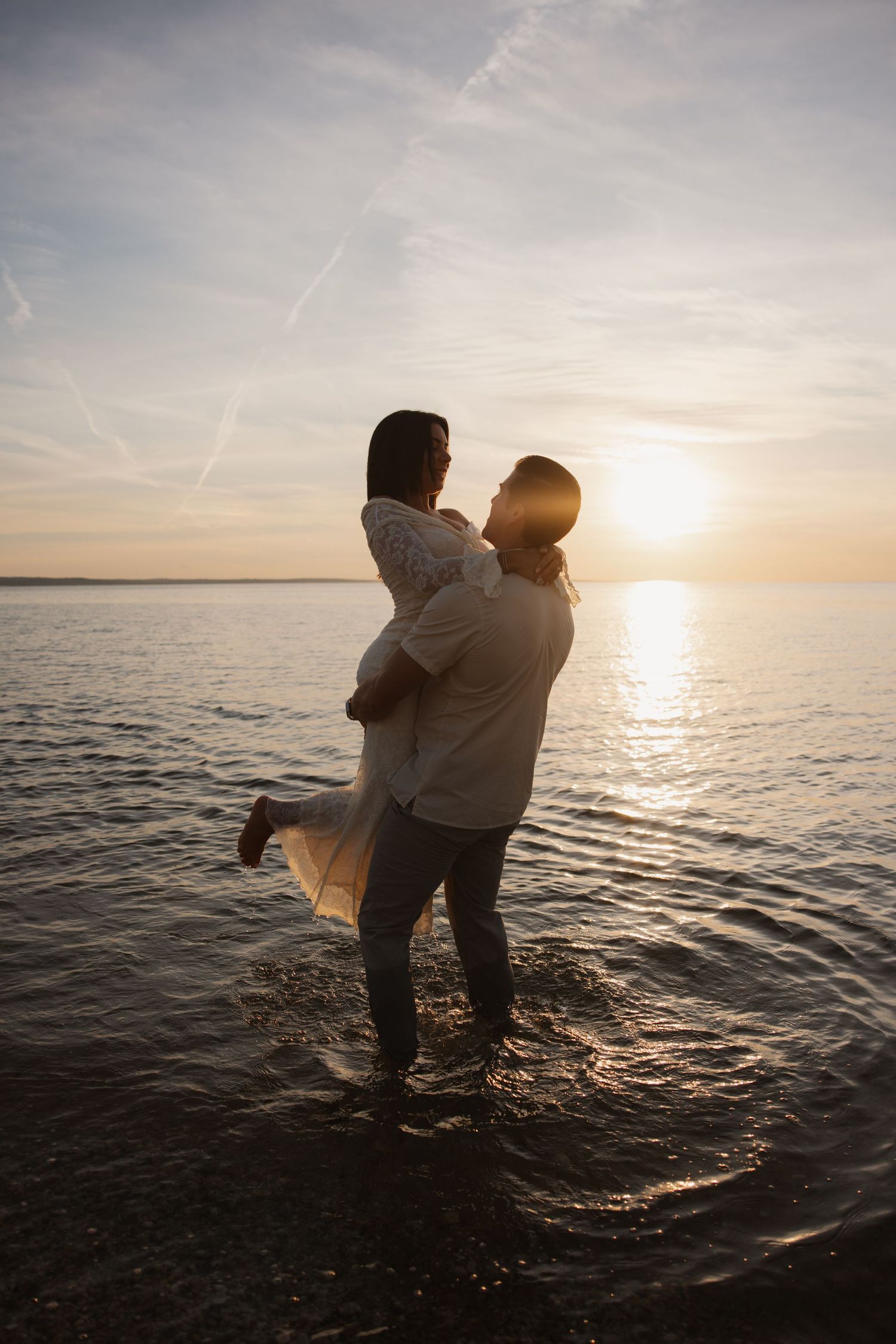 Couple embracing at sunset on a beach, standing in shallow water with golden light reflecting on the ocean.