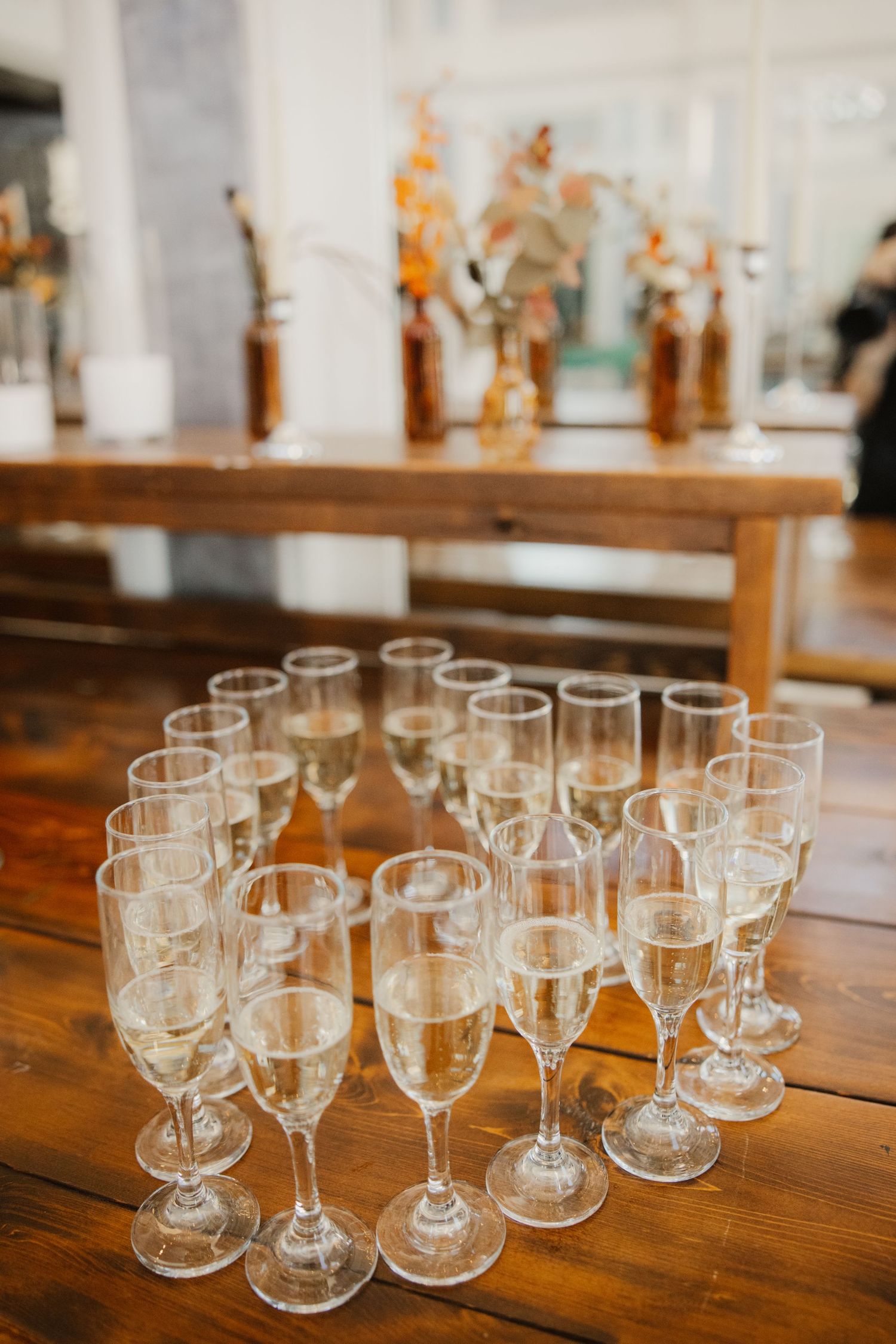 Glasses of champagne arranged on a rustic wooden table at a wedding reception with amber bottles and autumn flowers in the background.
