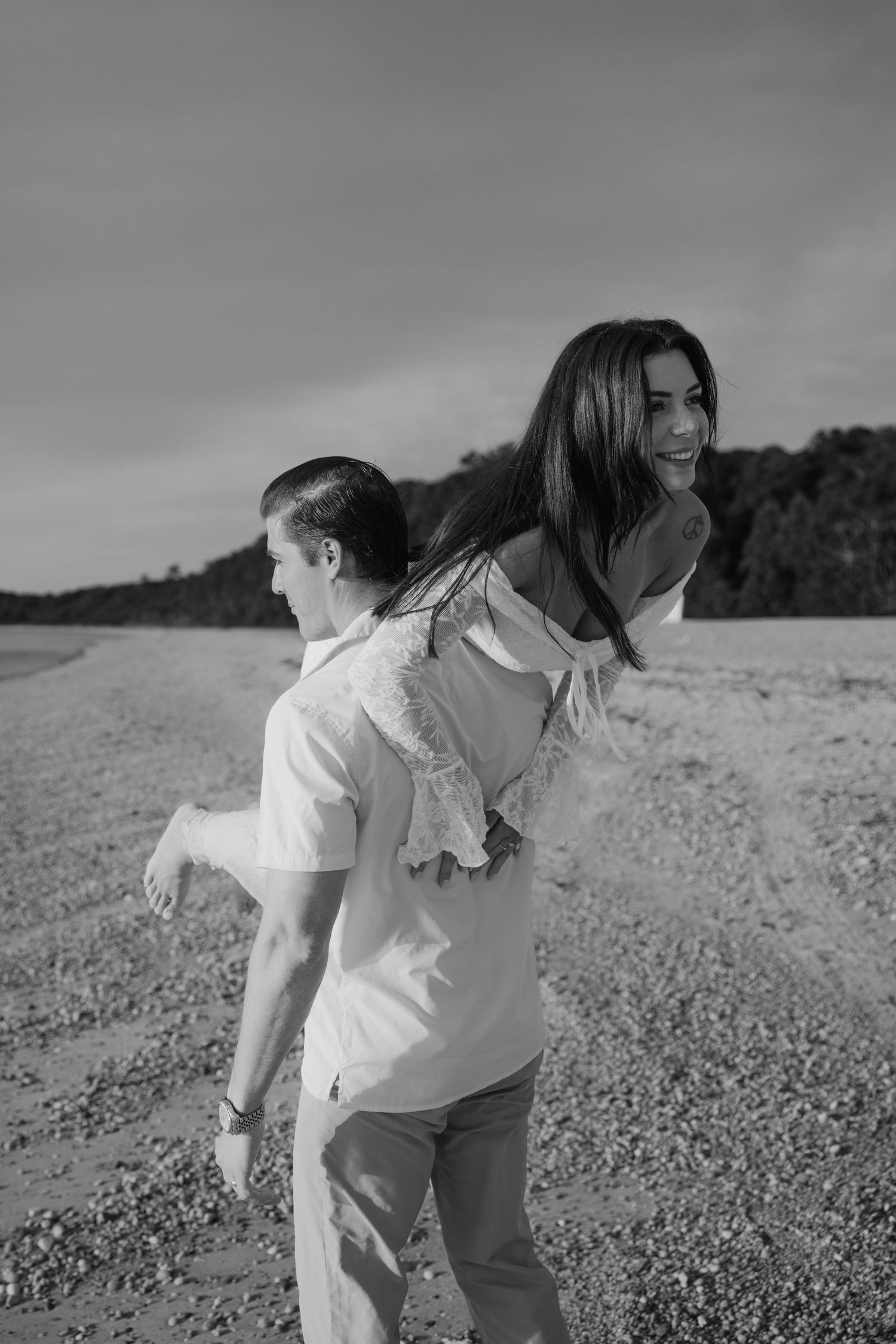 Black and white photo of a man giving a smiling woman a piggyback ride on a sandy beach.