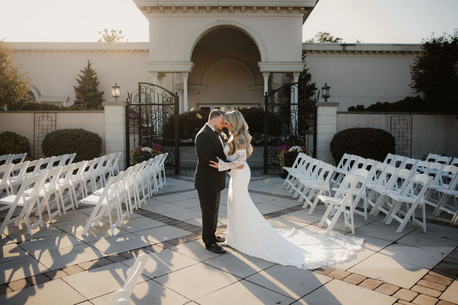 Bride and groom share a romantic kiss on a sunlit outdoor ceremony patio with white chairs.