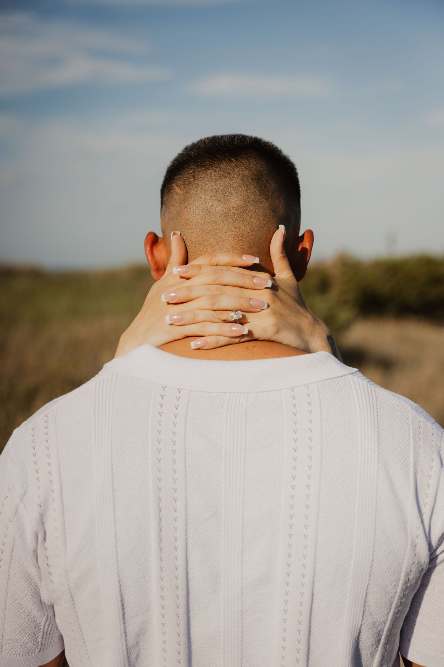 Close-up of woman's hand with engagement ring resting on back of man's neck outdoors.