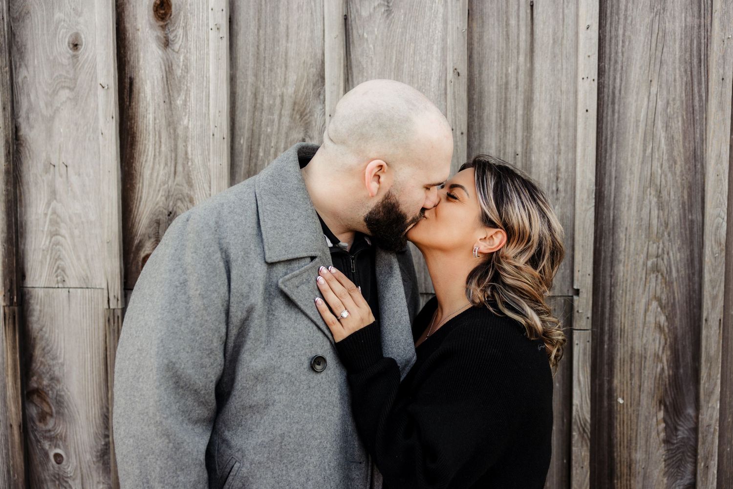 A couple shares a romantic kiss against a rustic wooden fence wall.