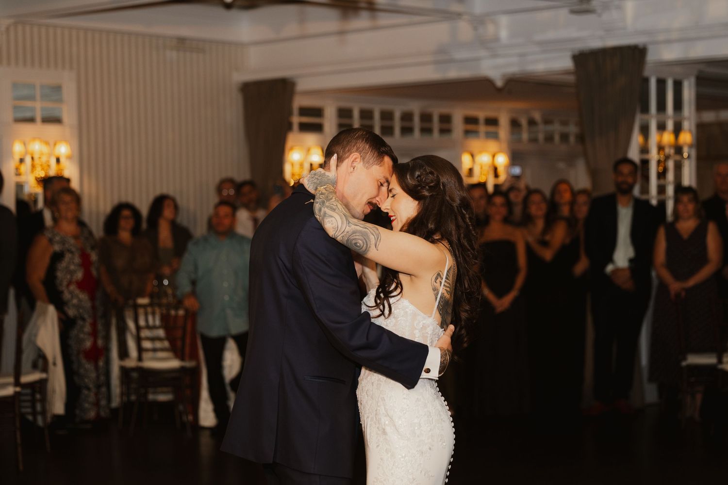 Bride in a lace gown and groom in a dark suit share a first dance as wedding guests watch in an elegant ballroom.
