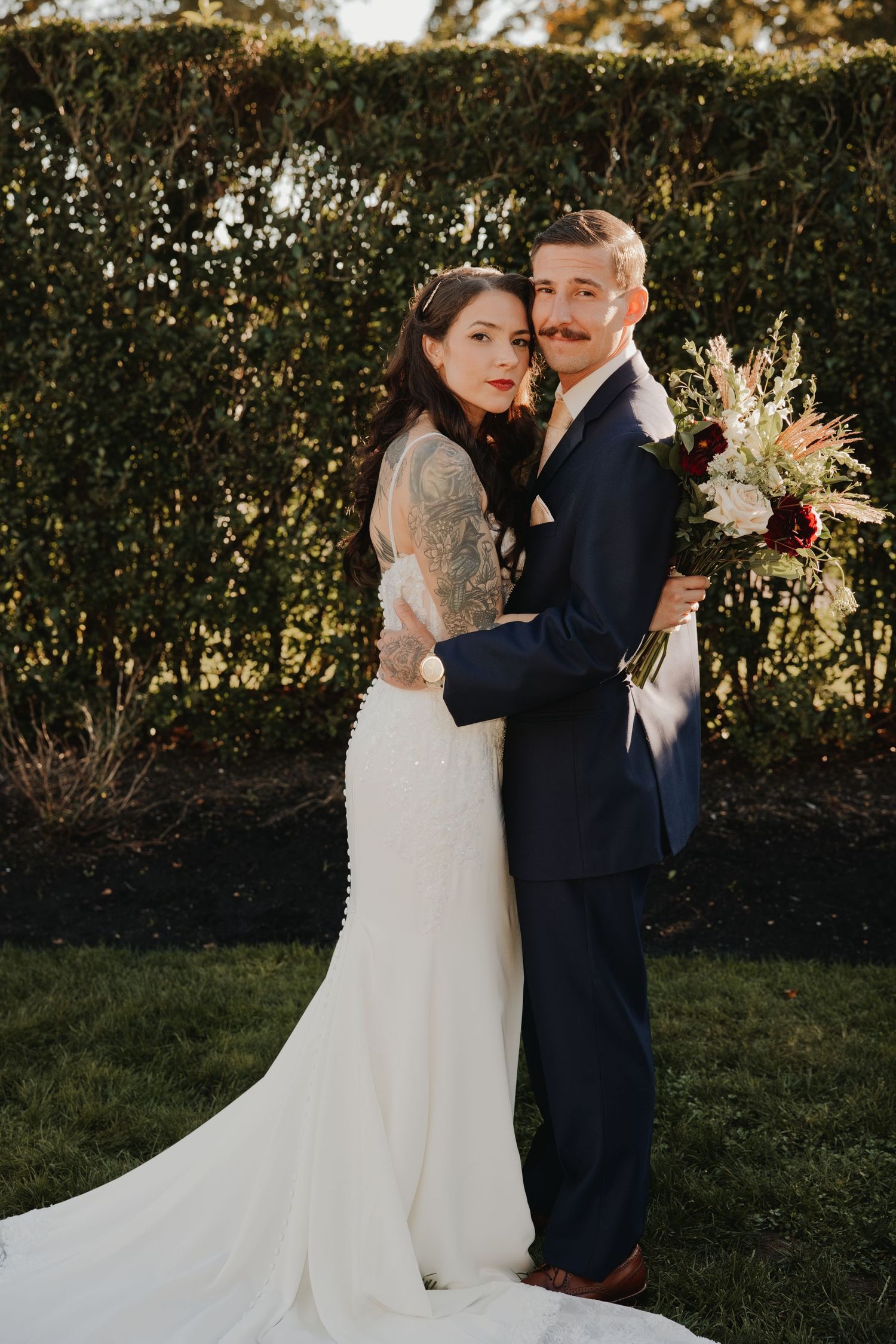 Bride in a white lace gown and groom in a navy suit embrace in front of a green hedge, holding a burgundy and blush bouquet.