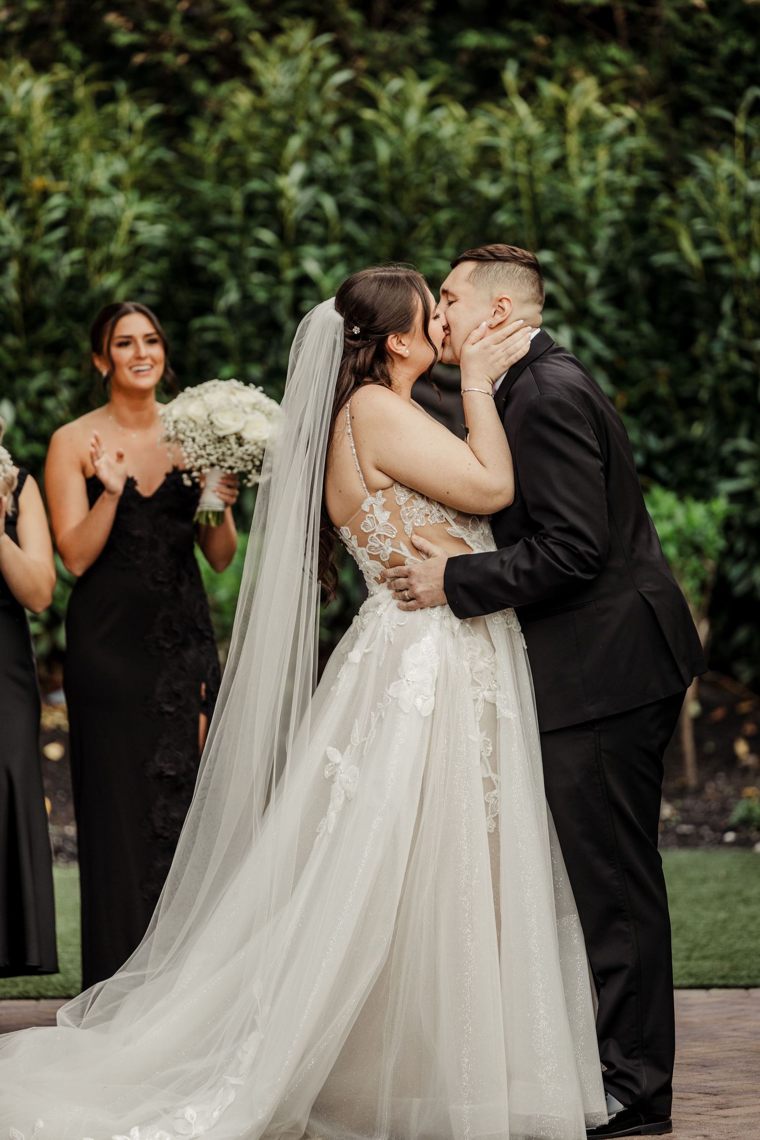 Bride and groom share their first kiss at outdoor wedding ceremony with bridesmaids in black gowns.