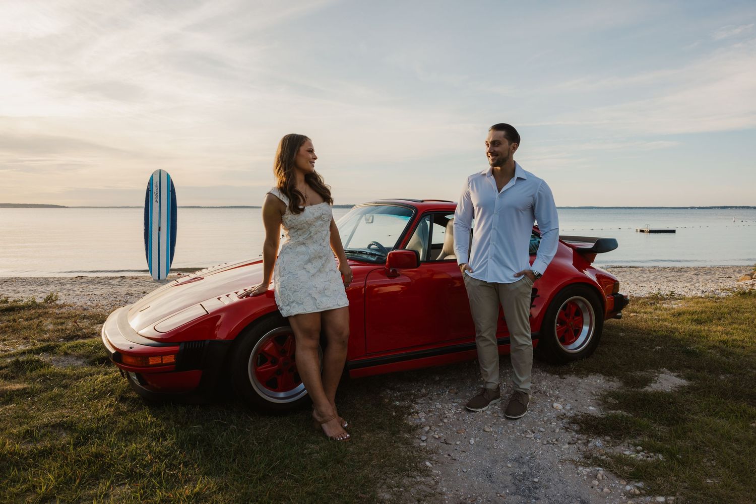 Couple smiling beside a red Porsche 911 near a beach with a blue surfboard at sunset.