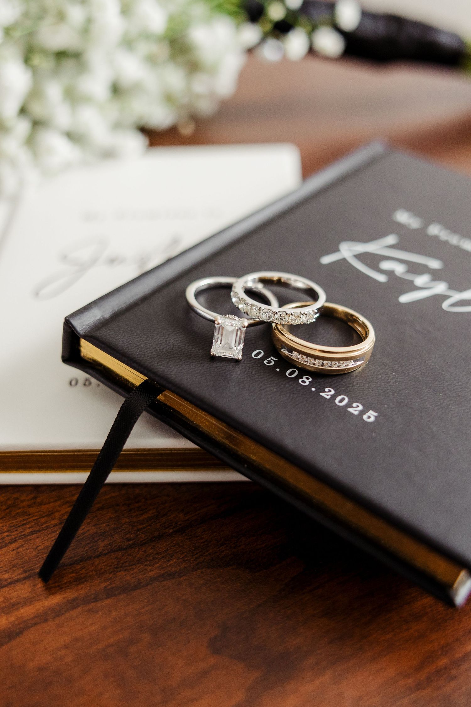 Three wedding rings rest on a black vow book dated 05.09.2025 on a wooden table.