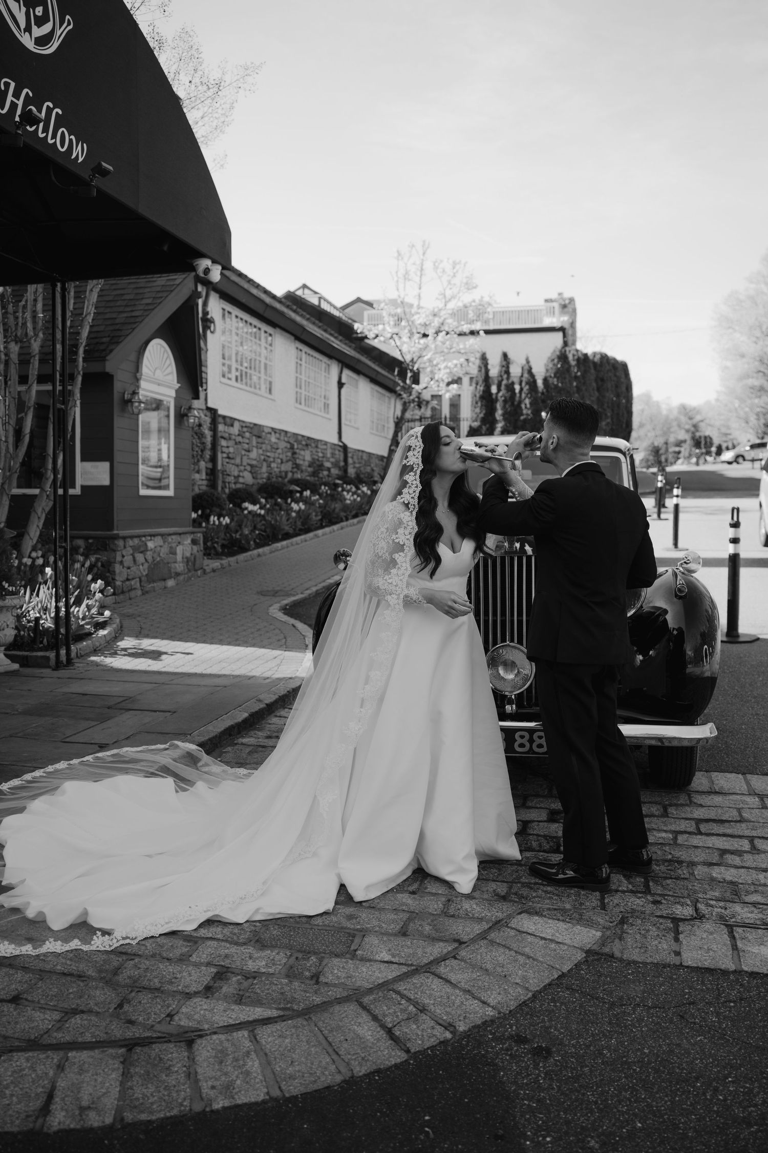 Black and white photo of a bride and groom posing beside a vintage car on a cobblestone street outside an elegant venue.