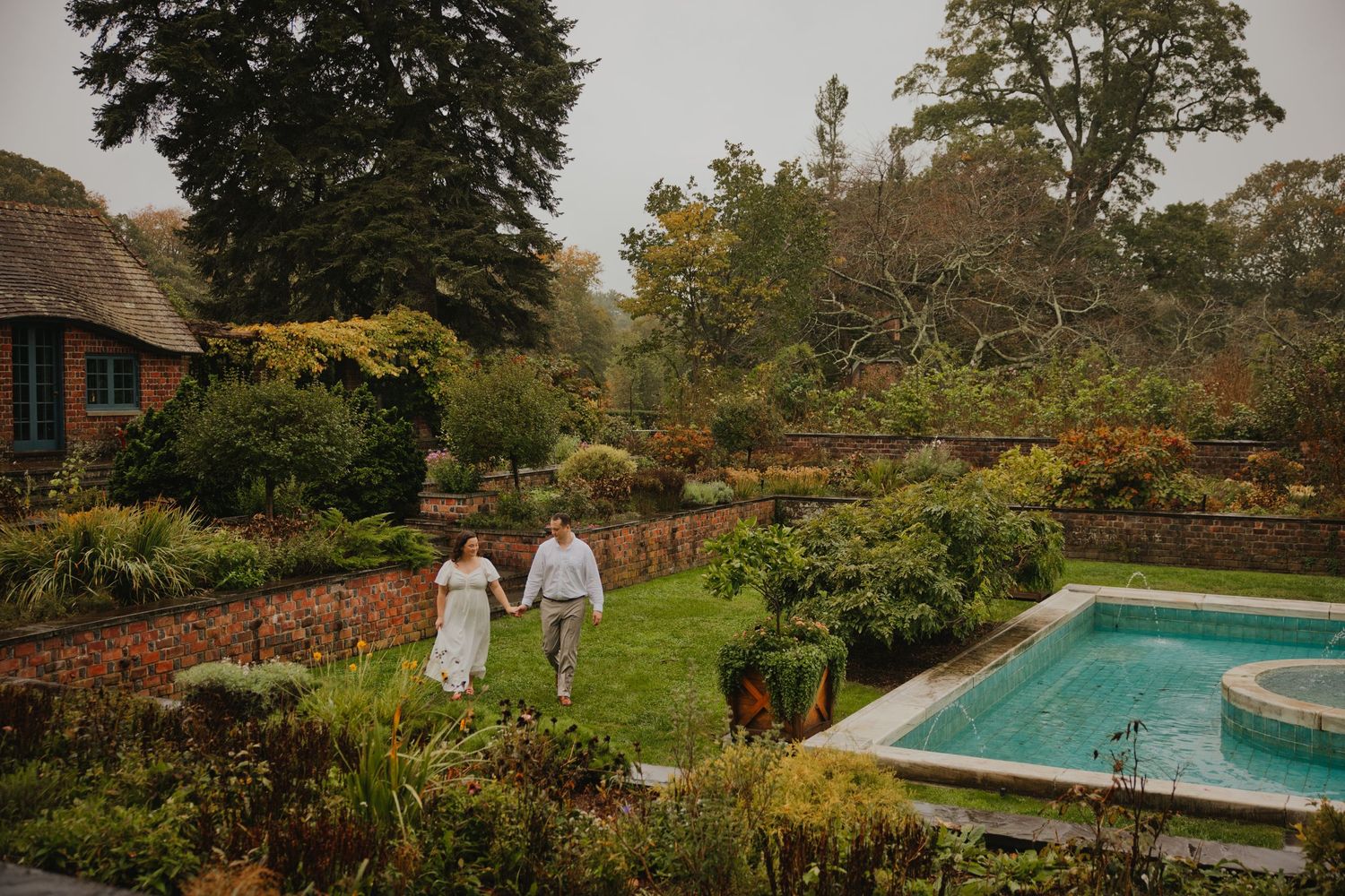 Couple holding hands walking through a lush garden estate with a pool on a misty autumn day.