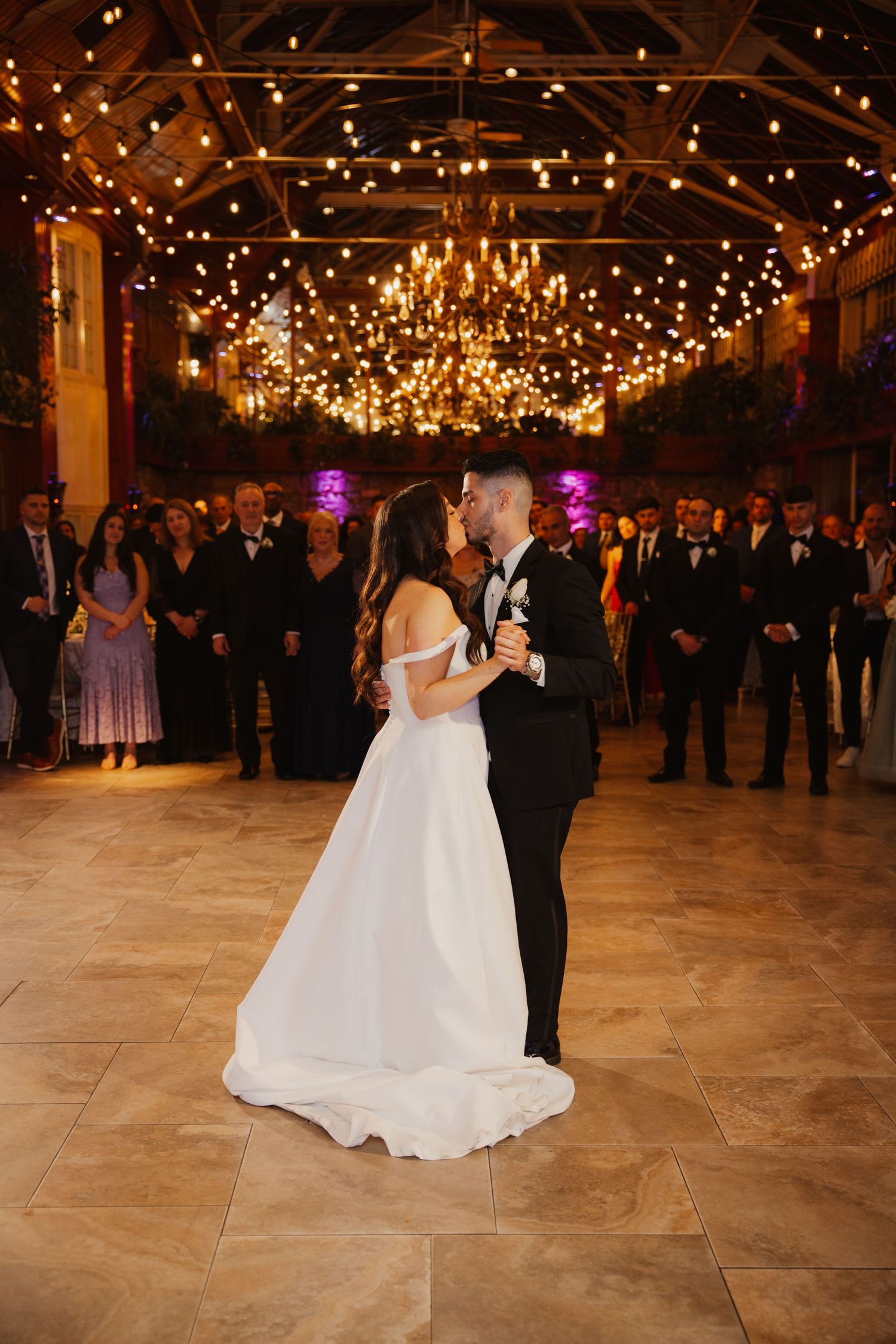 Bride and groom share first kiss on dance floor under glowing chandeliers and string lights at reception.