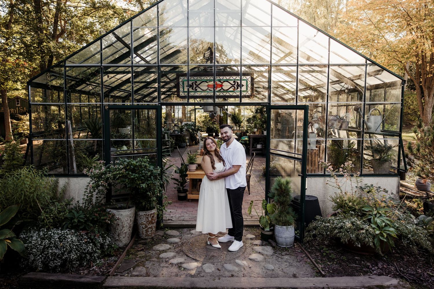 A couple poses together in front of a beautiful glass greenhouse surrounded by plants.