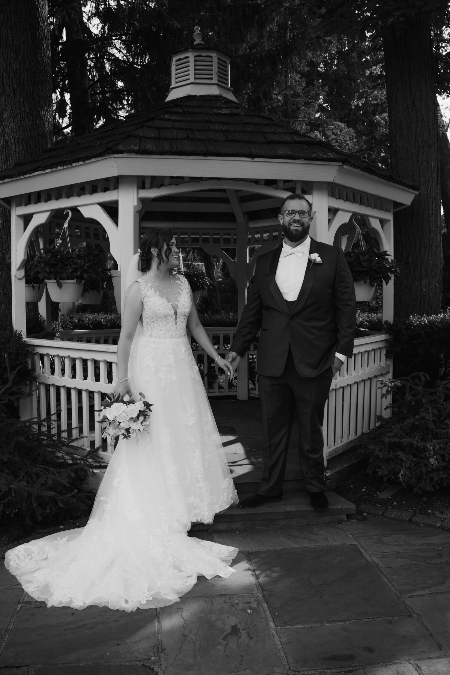 Black and white photo of bride and groom holding hands before a white gazebo on their wedding day.