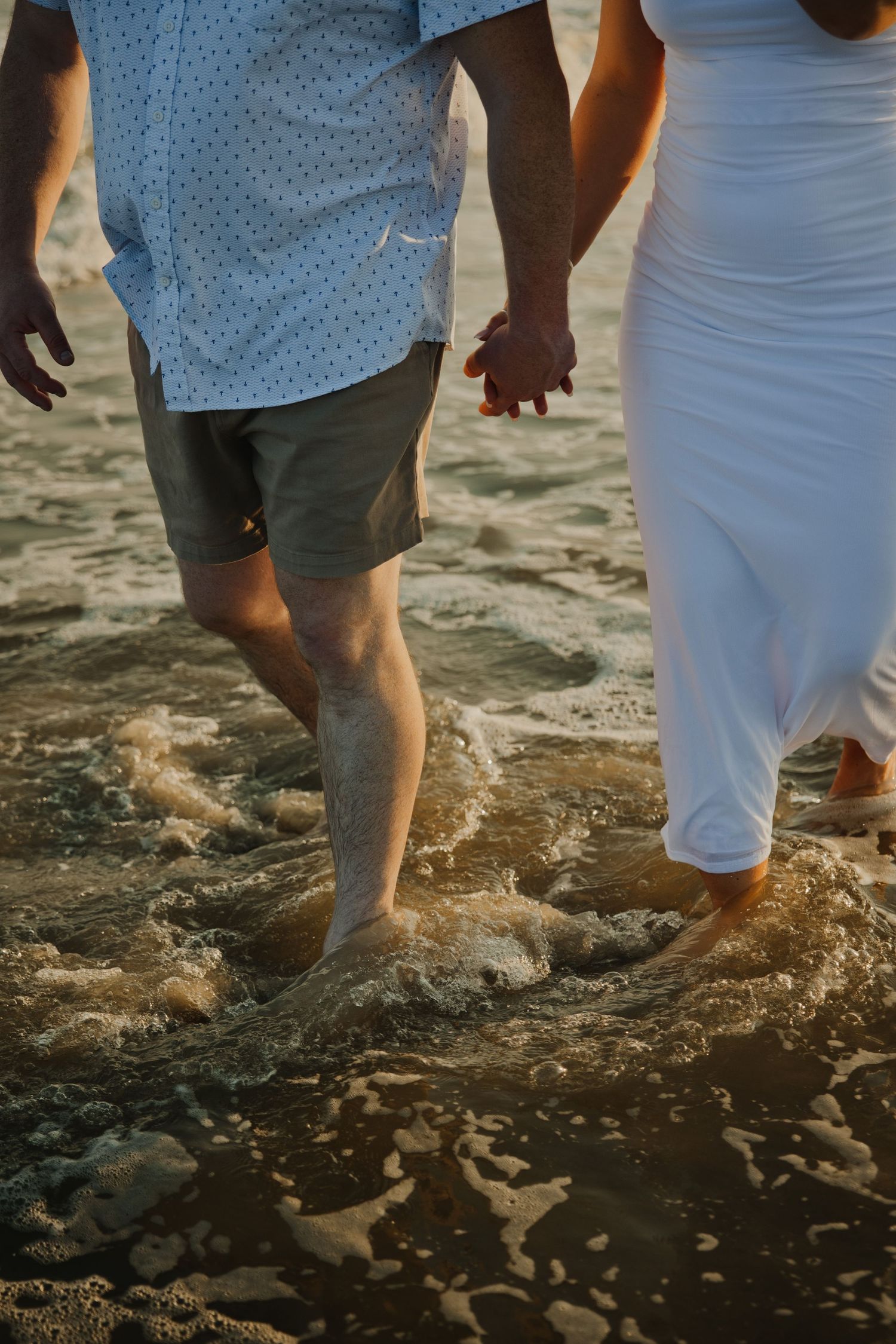 Close-up of couple holding hands while walking barefoot through ocean waves on sandy beach.