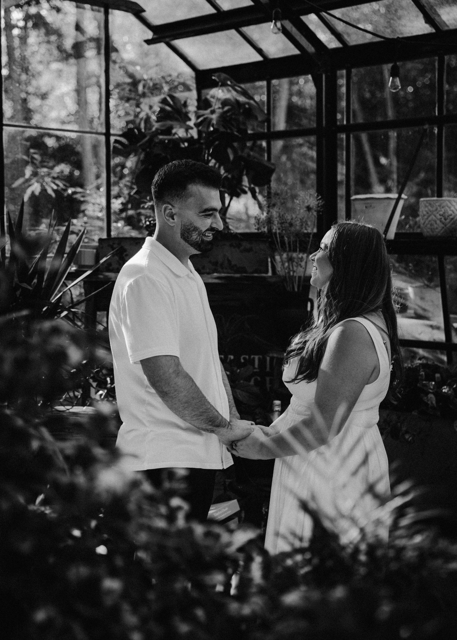 A couple holds hands and smiles at each other inside a greenhouse in black and white.