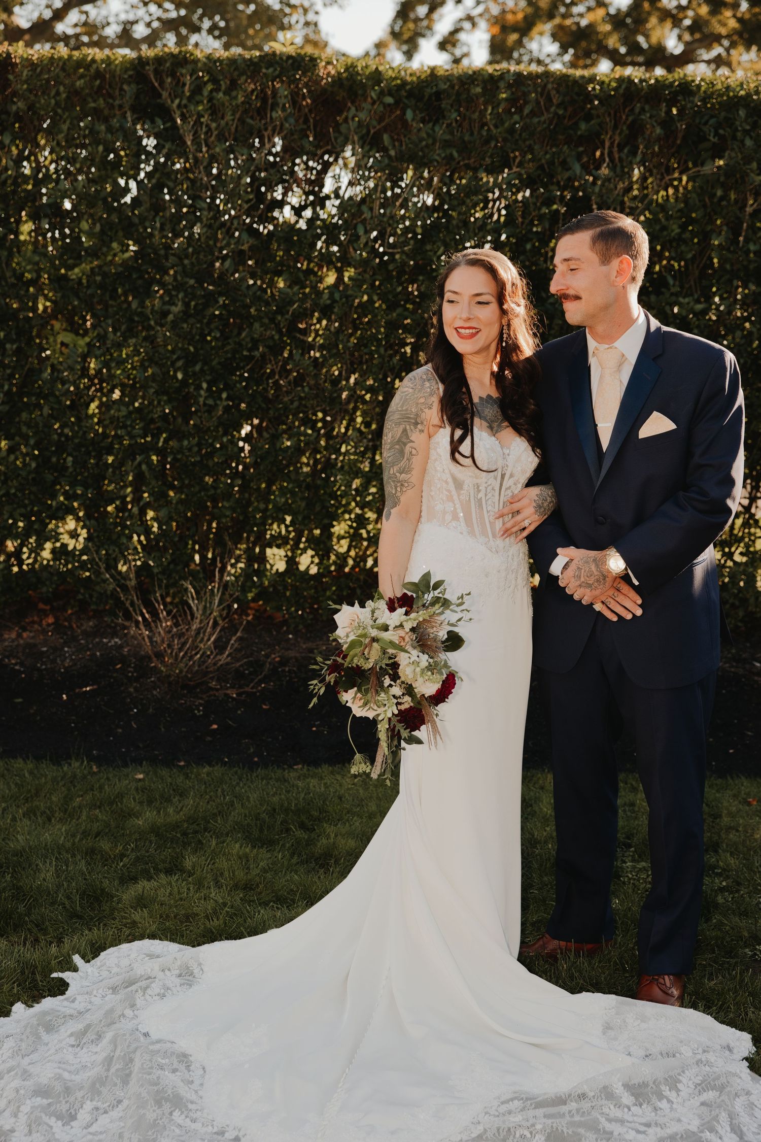 Smiling bride in a lace mermaid wedding gown holding a floral bouquet stands beside a groom in a navy suit on a lawn by a green hedge.