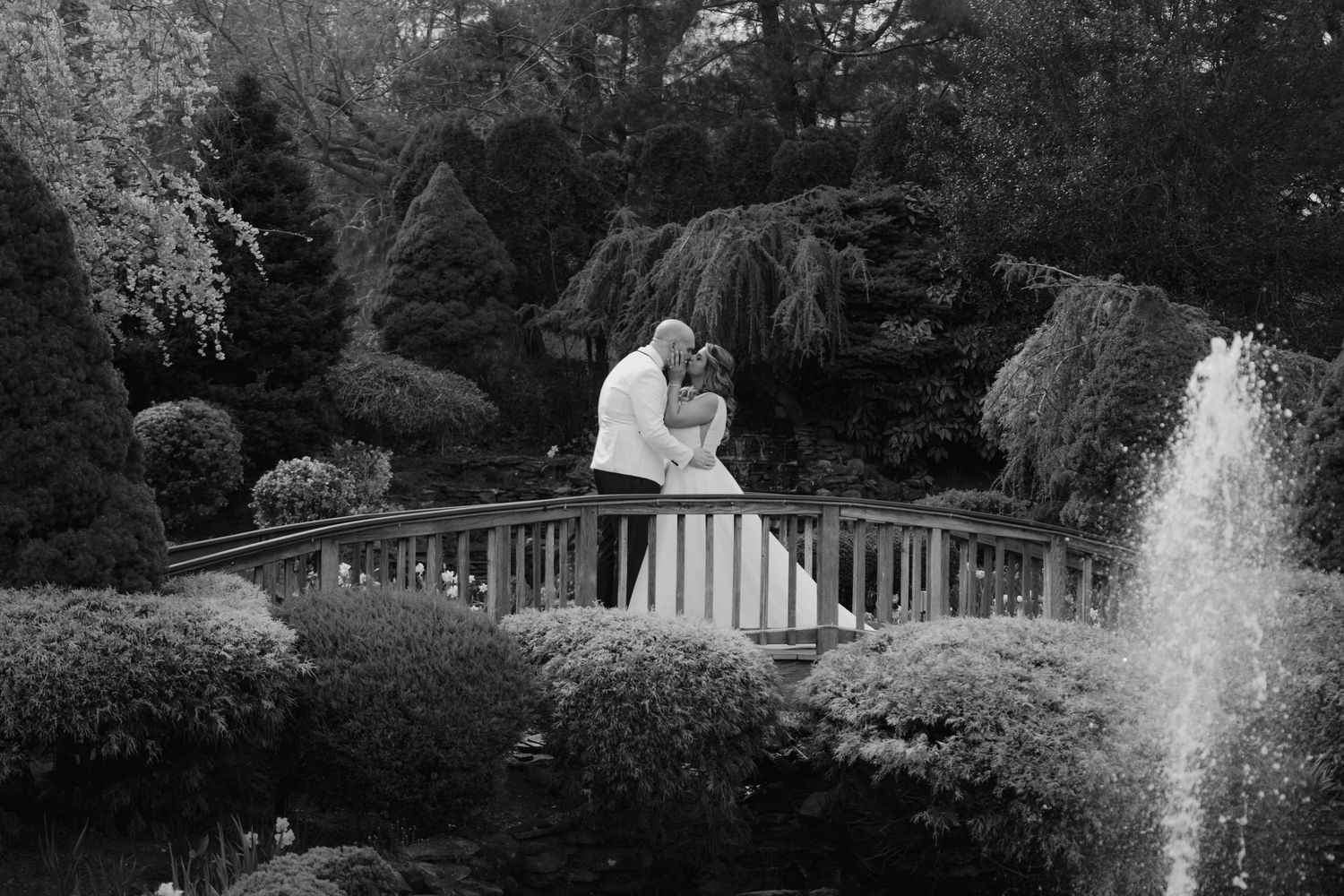 Black and white photo of bride and groom embracing on garden bridge near fountain.