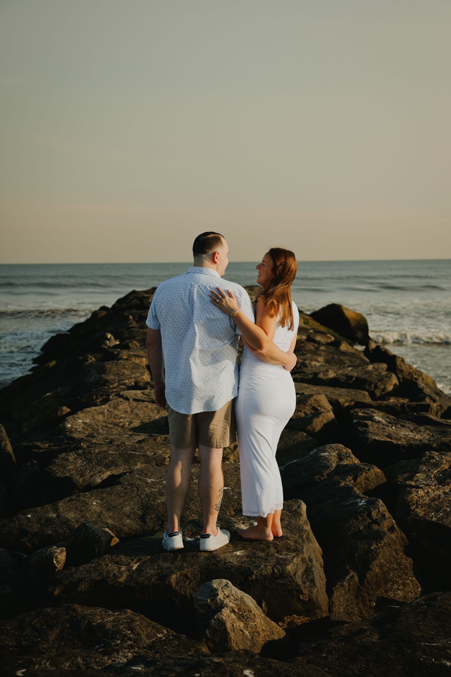 Couple standing on rocky jetty embracing while gazing at ocean waves during golden hour sunset.