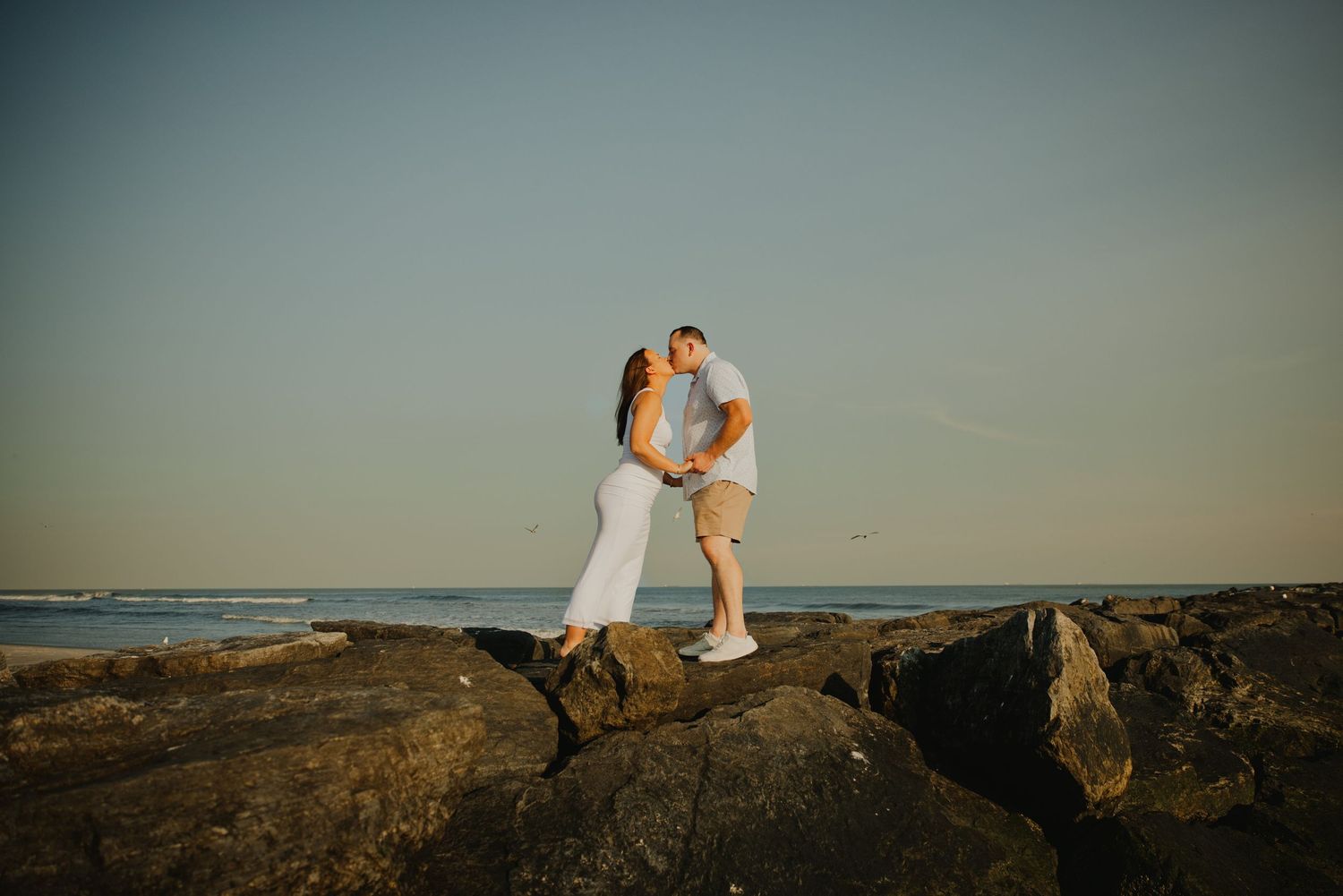 Couple kisses on rocky shoreline at sunset with calm ocean and clear blue sky in background.