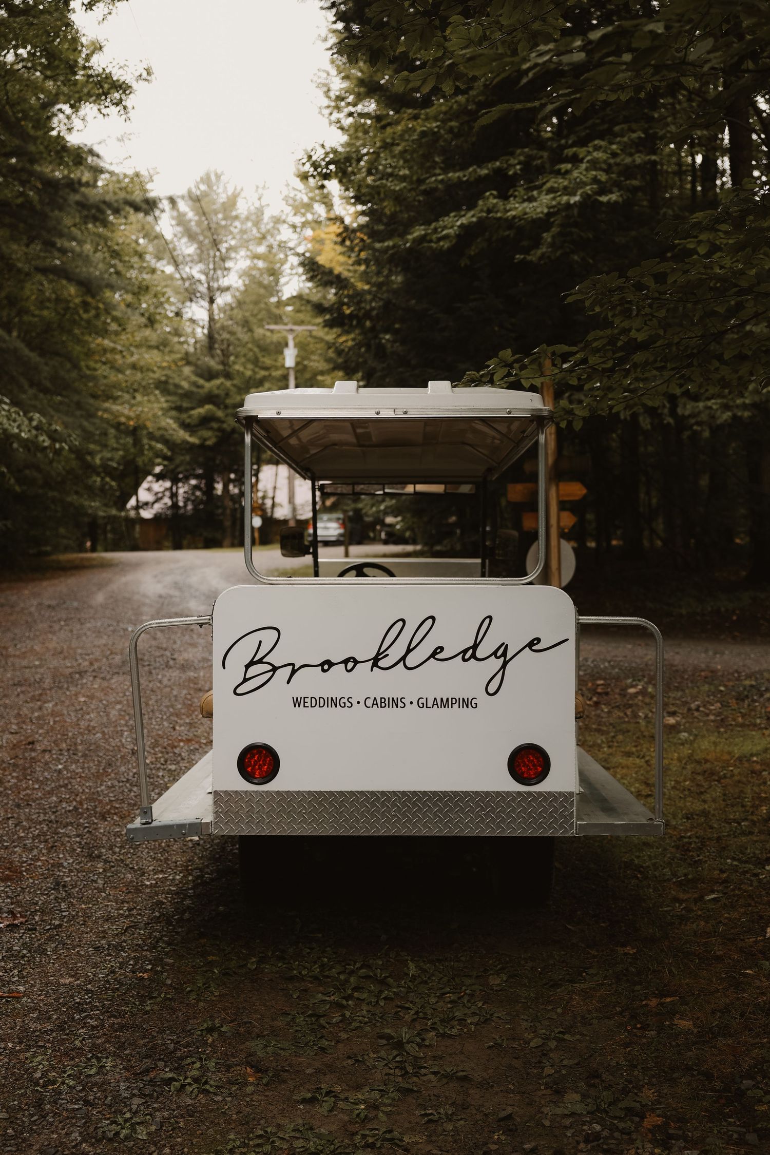 Brookledge wedding venue golf cart shuttle parked on wooded gravel path among tall pine trees.