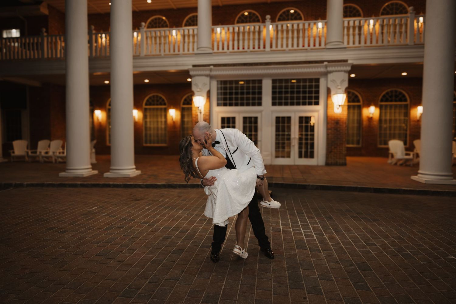 Groom dips bride in romantic pose under columned venue portico in evening wedding portrait.