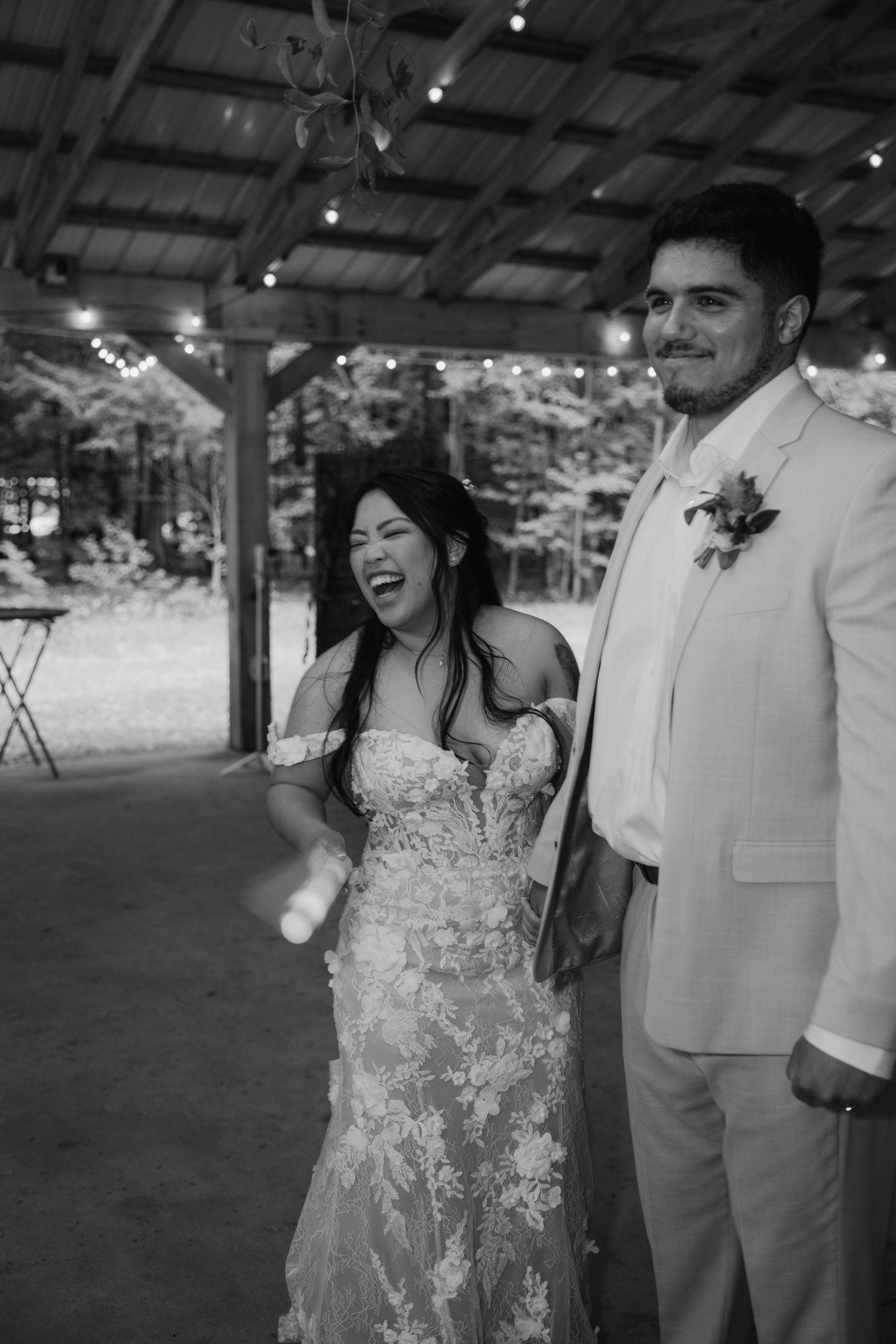 Black and white photo of a laughing bride in lace gown and groom in white suit at an outdoor wedding venue.