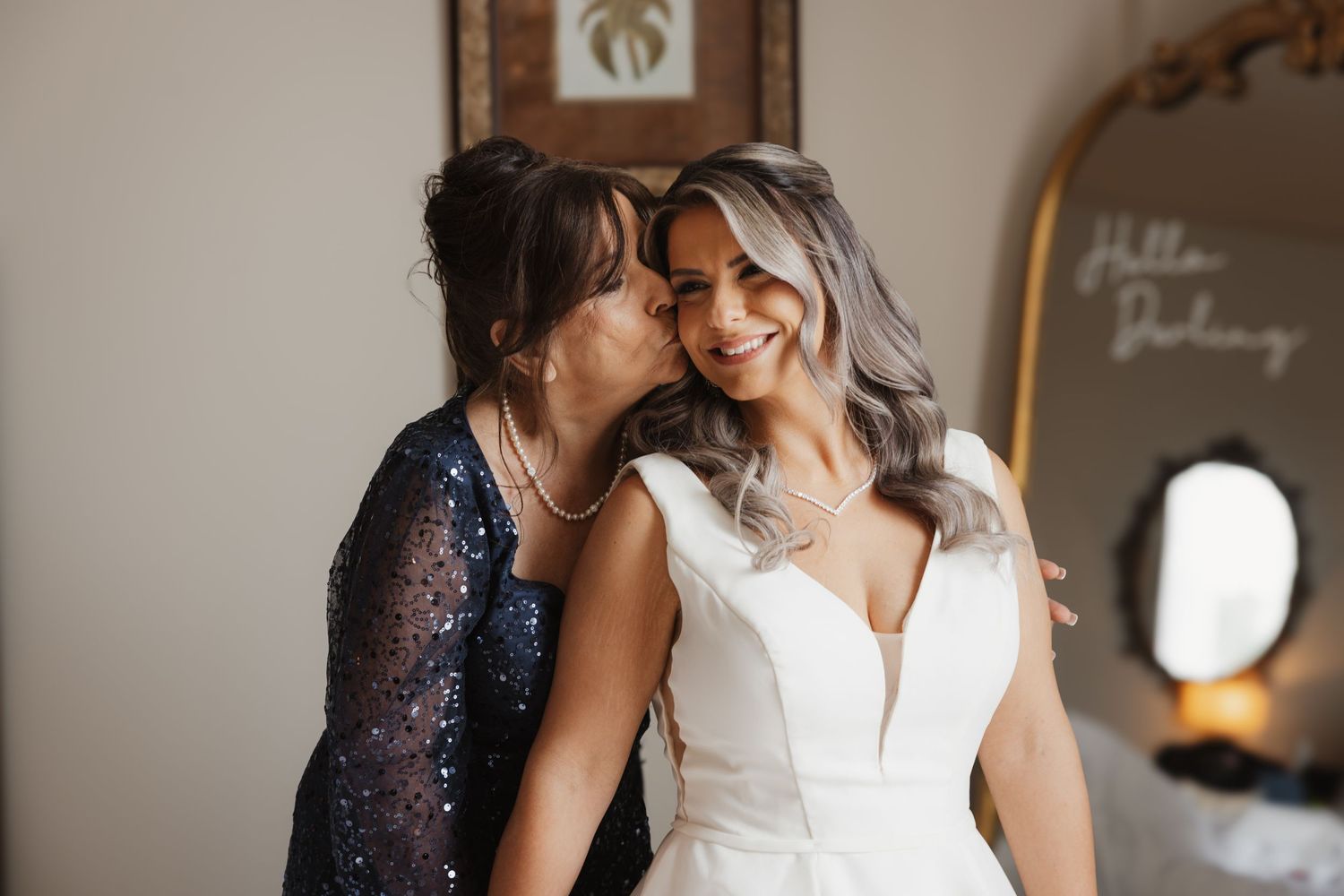 Joyful mother kisses smiling bride in white wedding gown in front of Hello Darling mirror sign.