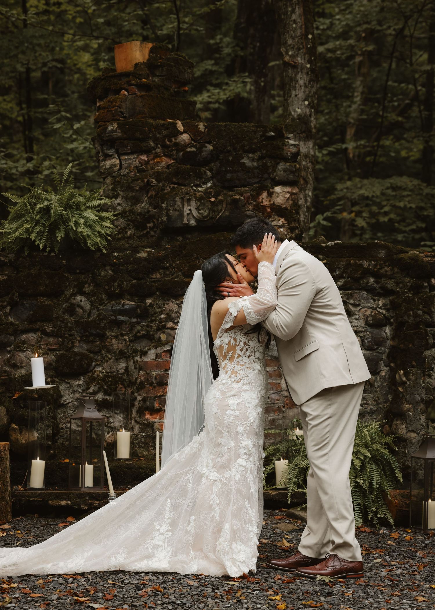 Bride and groom share first kiss at rustic stone altar surrounded by candles in a lush forest setting.