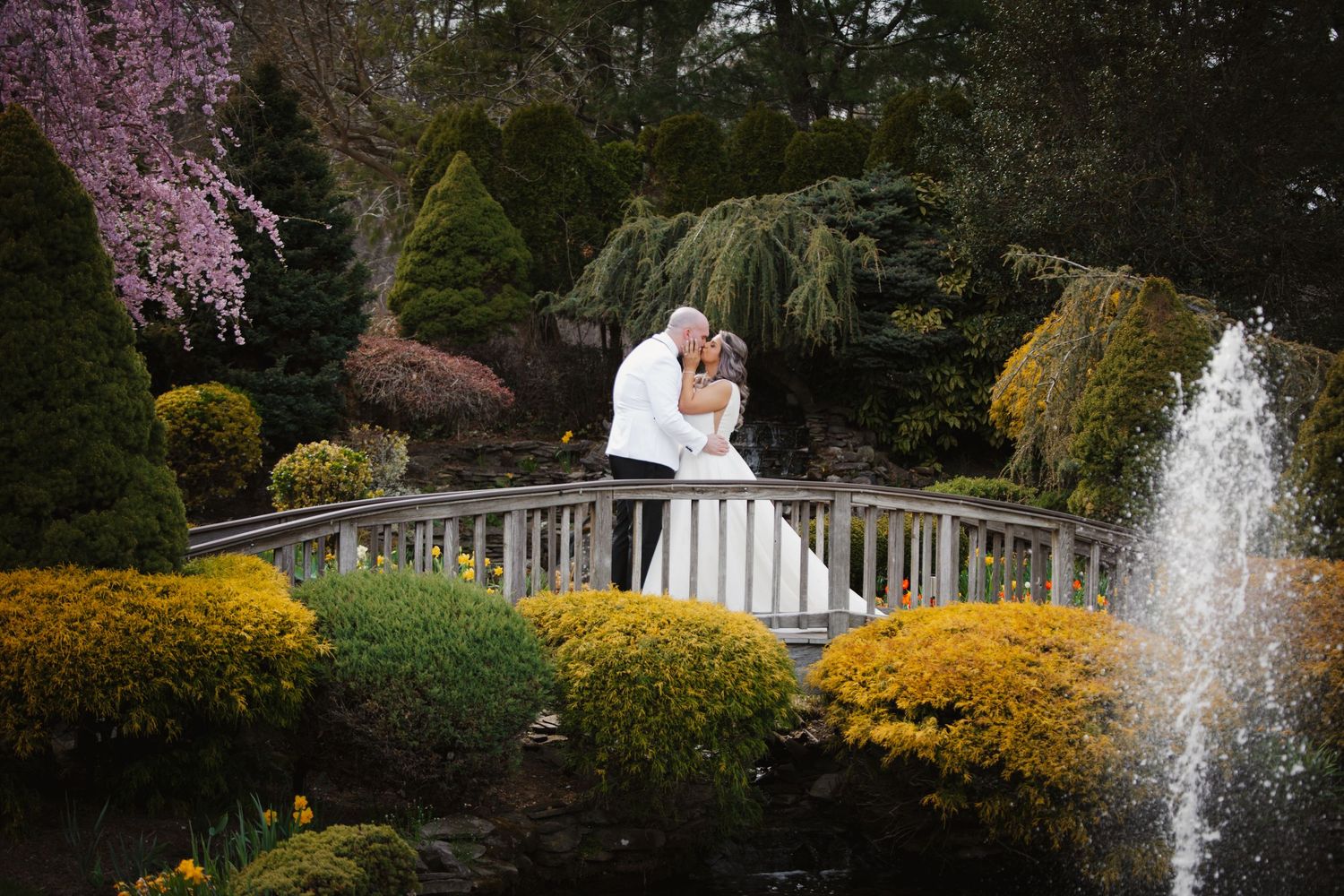 Bride and groom kissing on a white bridge surrounded by colorful gardens and a fountain at an outdoor wedding venue.