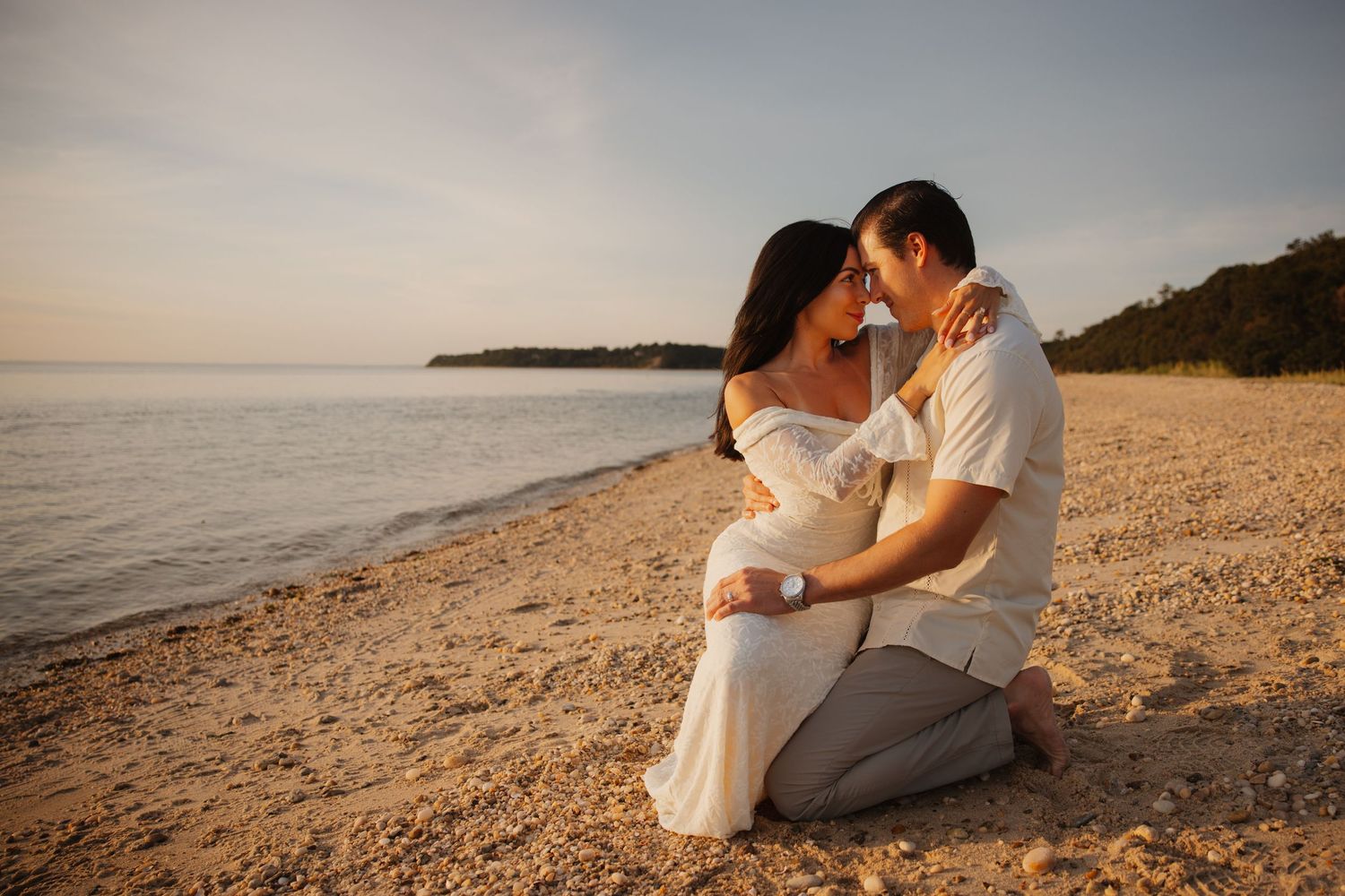 Couple sharing a romantic moment on a sandy beach at golden hour, woman in white dress, man in beige outfit.